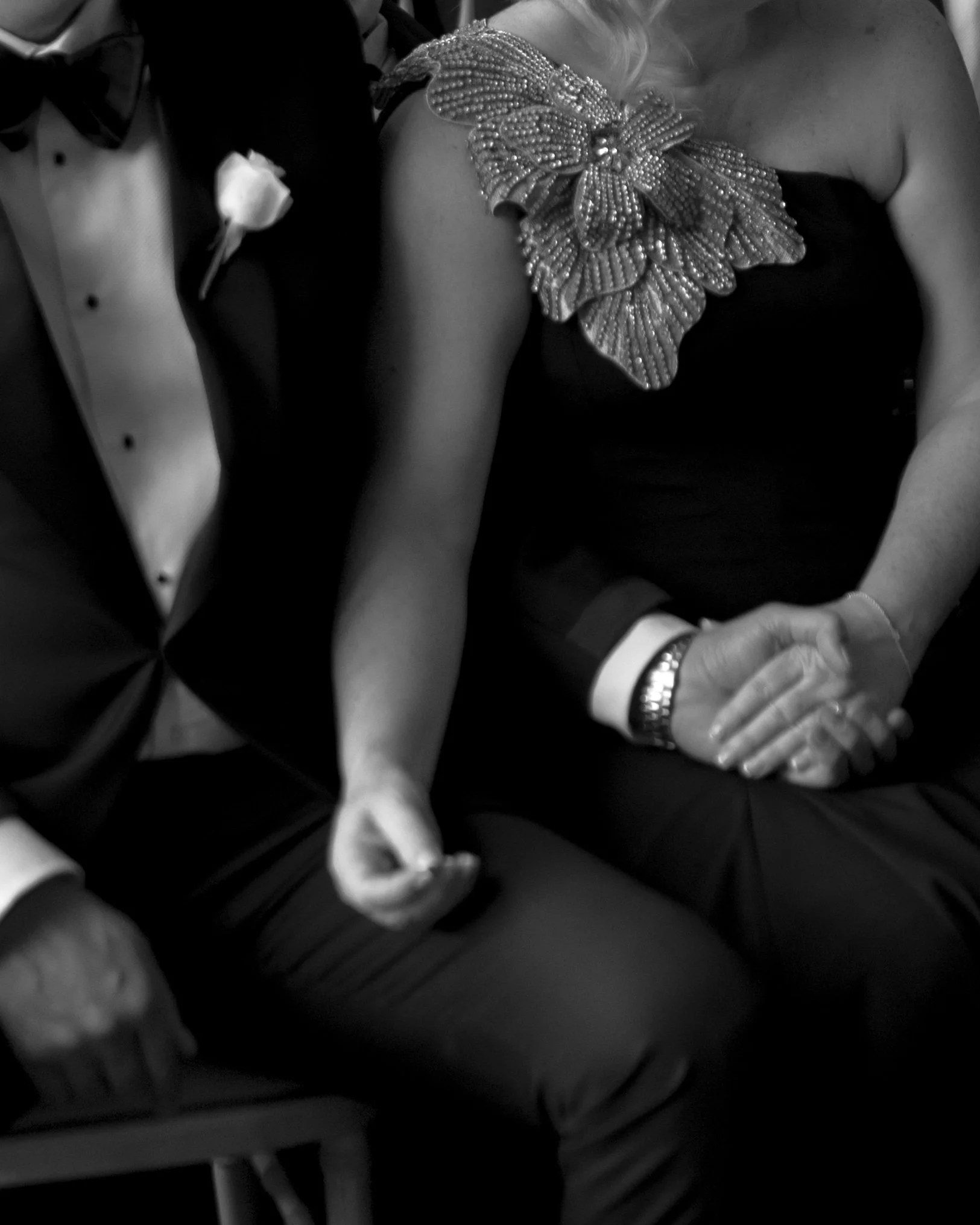 Close-up of a man in a tuxedo and a woman in a black dress, holding hands at a black tie wedding ceremony.