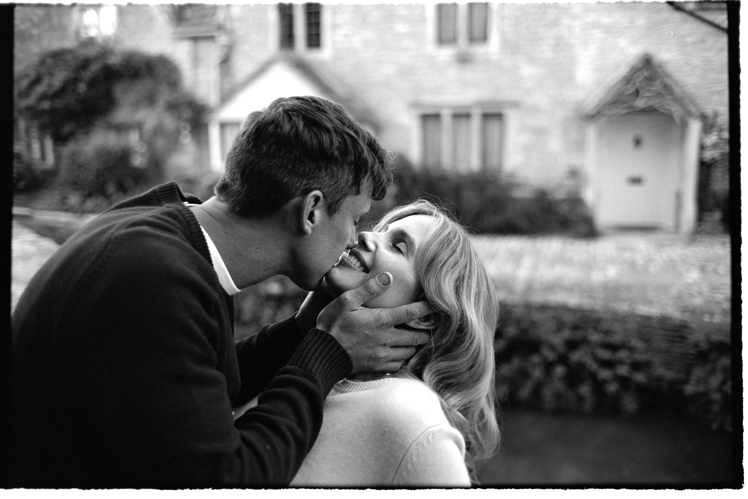 A black-and-white photo of a couple about to kiss outdoors, with the man holding the woman's face gently. The background shows a house with trees and bushes.