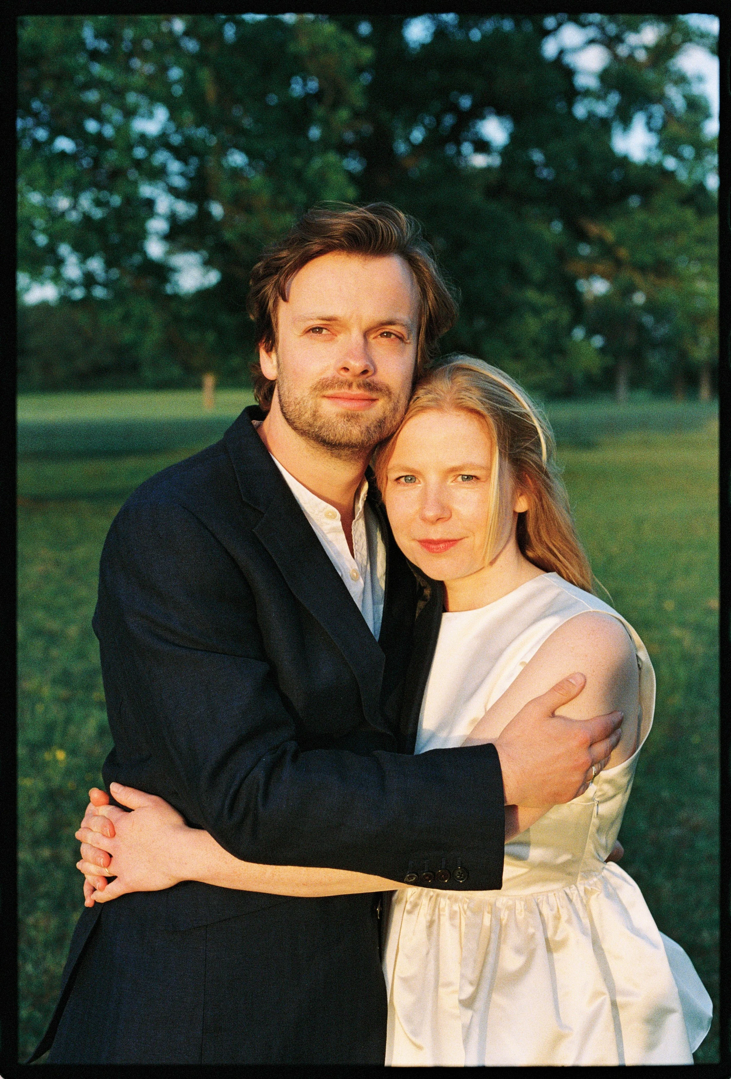 A man and a woman embracing outdoors during sunset, with trees and grass in the background.