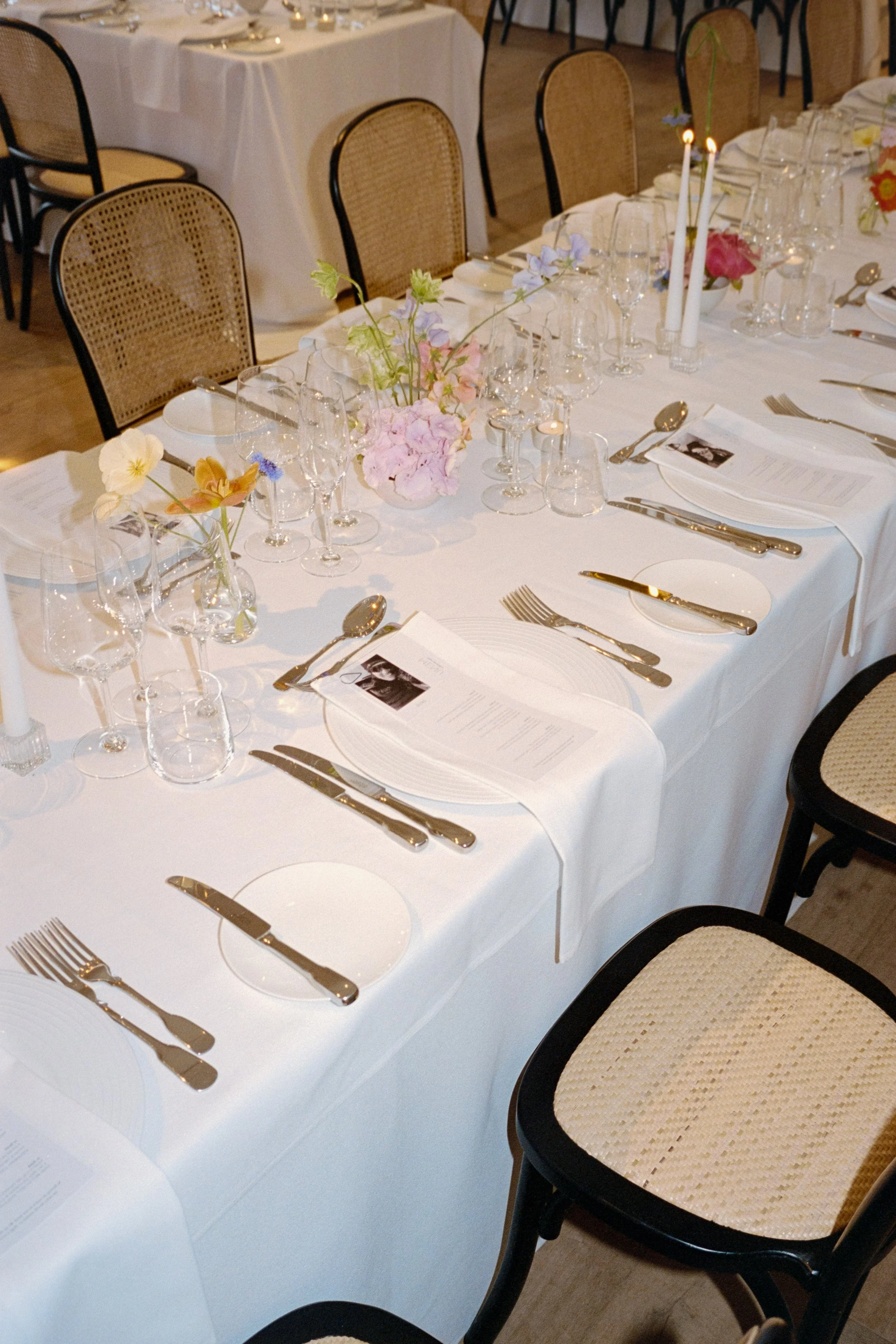 Elegant dining table set with white tablecloth, floral centerpieces, candles, and set-for-a-meal with glassware, plates, and silverware in a formal event setting.