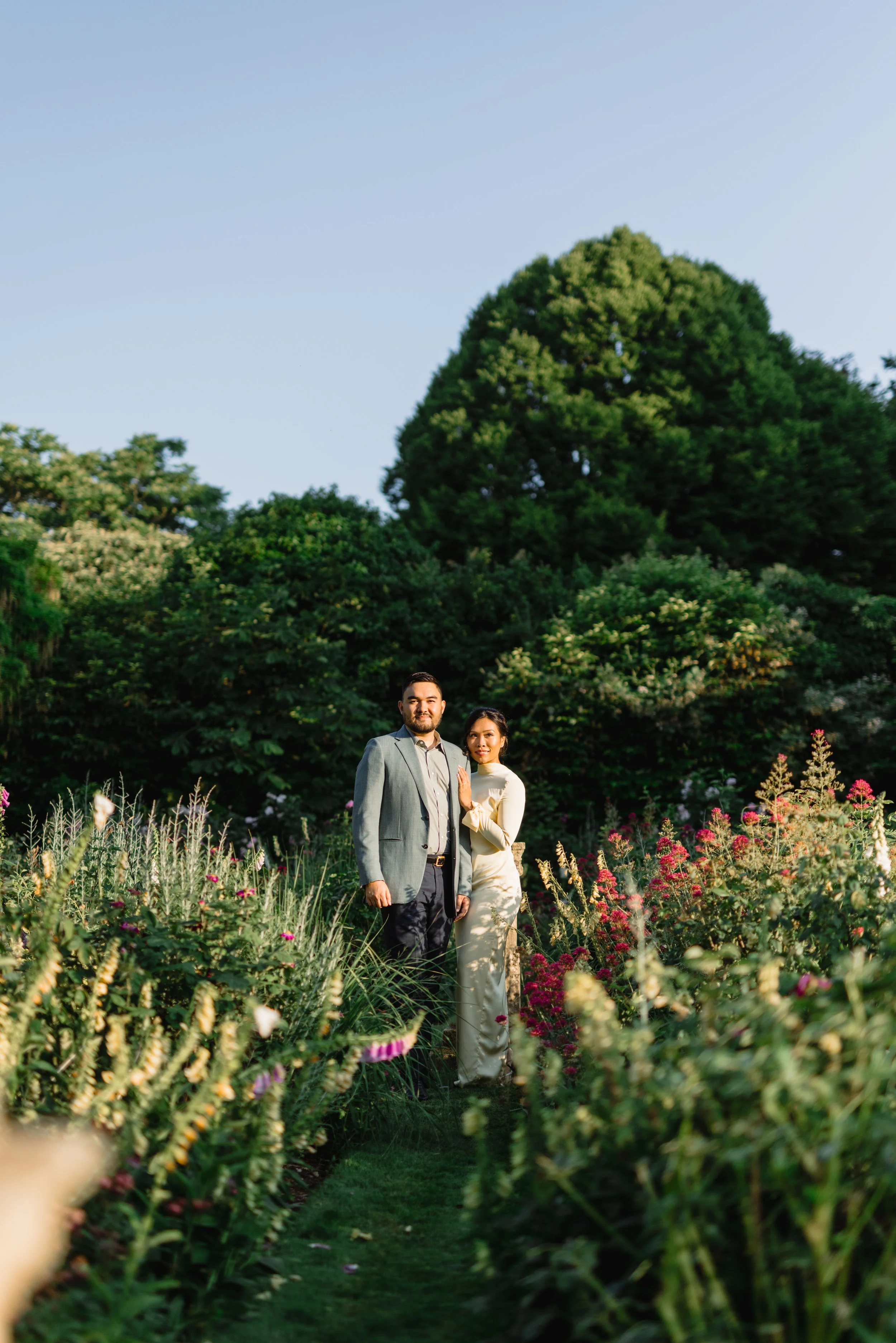 A couple standing among colorful flowers in a lush garden with large green trees in the background.