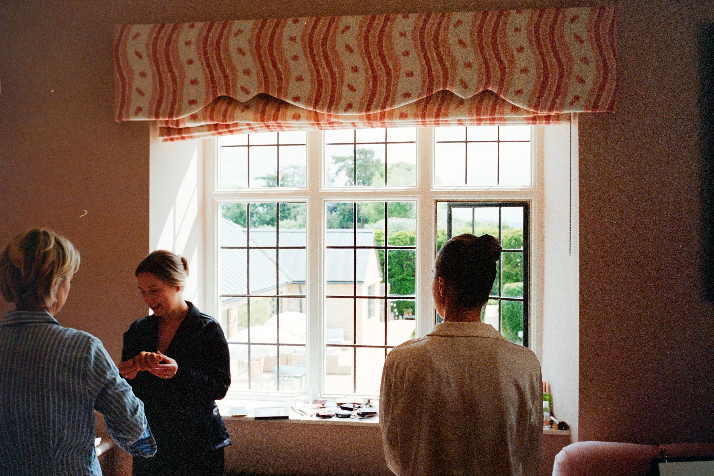 Three women standing by a large window with a patterned valance, engaging in conversation and exchanging small items in a brightly lit room.