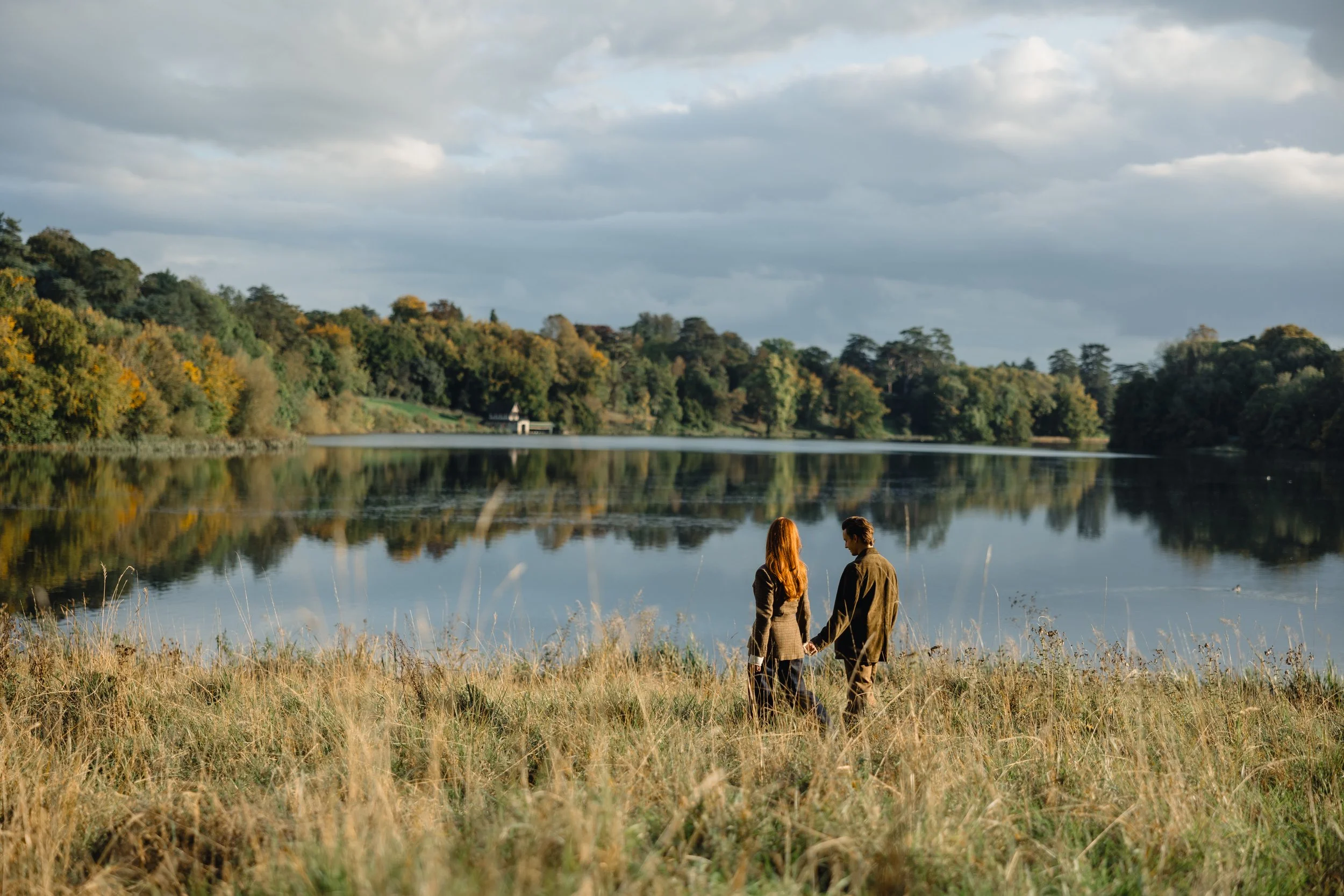 A couple holding hands standing in tall grass near a calm lake with a tree-covered shoreline in the background and a partly cloudy sky.