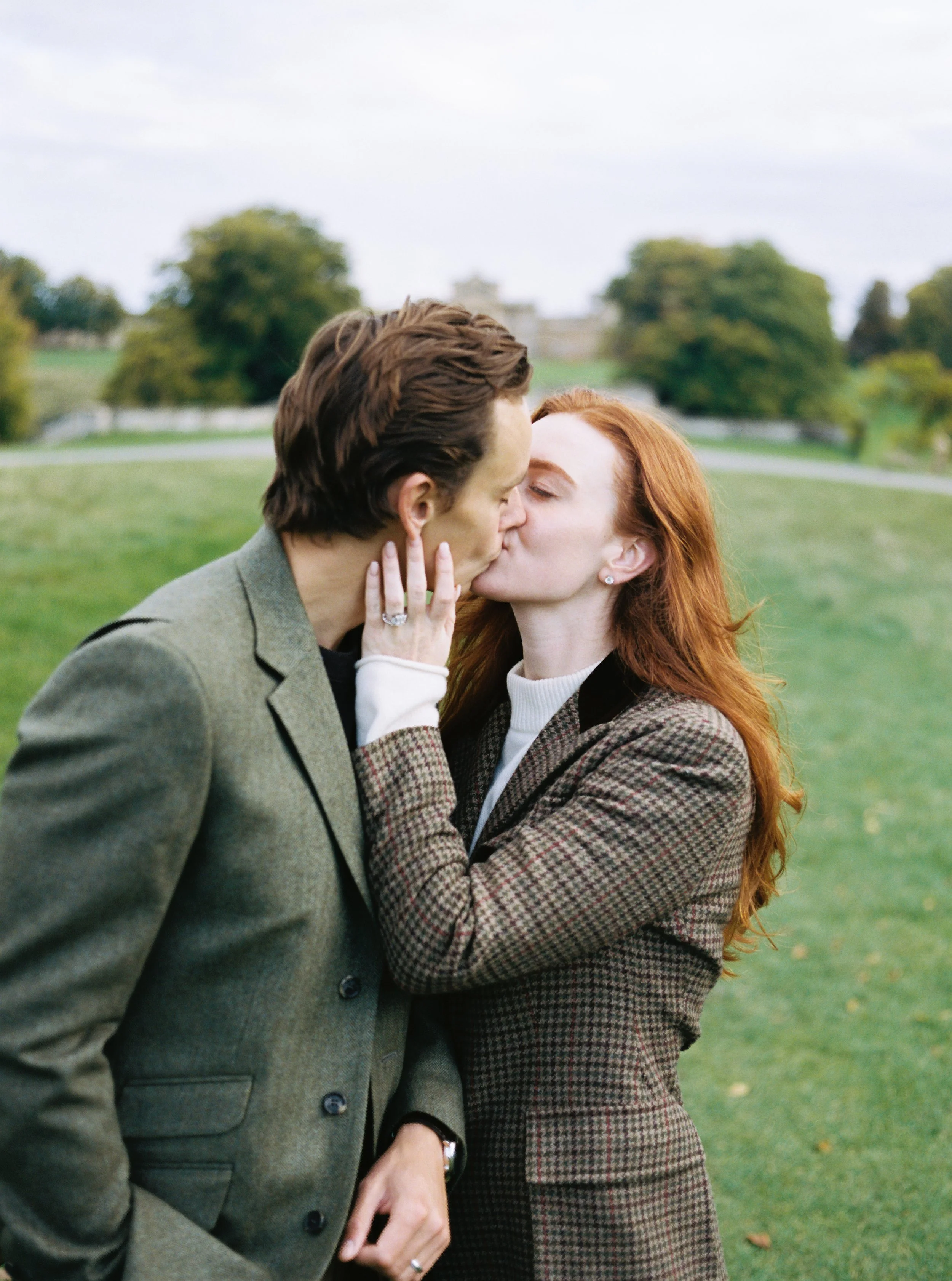 A man and woman kissing outdoors on a grassy area with trees and a cloudy sky in background.