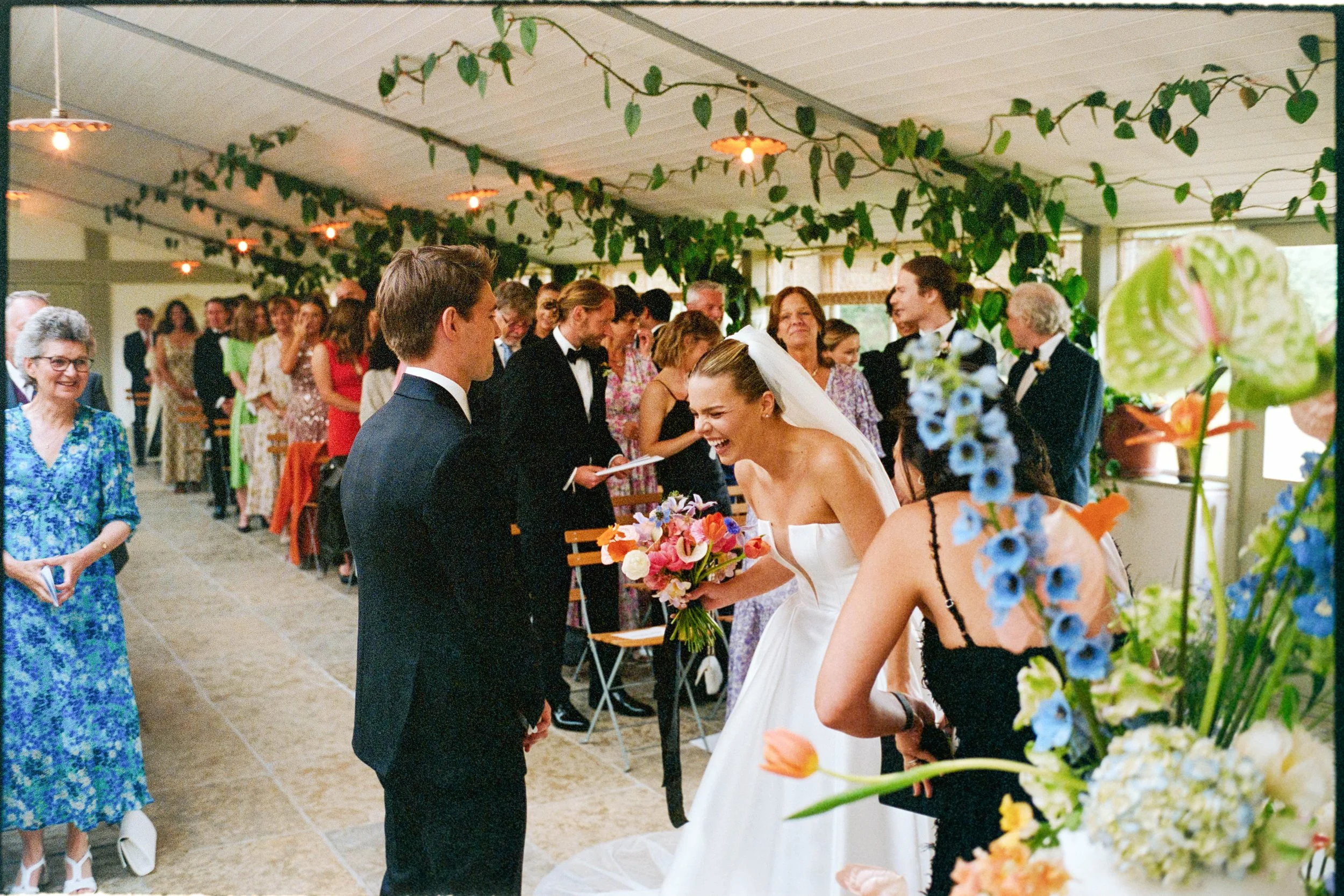 Bride in a wedding dress holding a bouquet of flowers, laughing and talking to a groom in a tuxedo, in a decorated indoor venue with green plants and hanging lights, surrounded by family and friends.