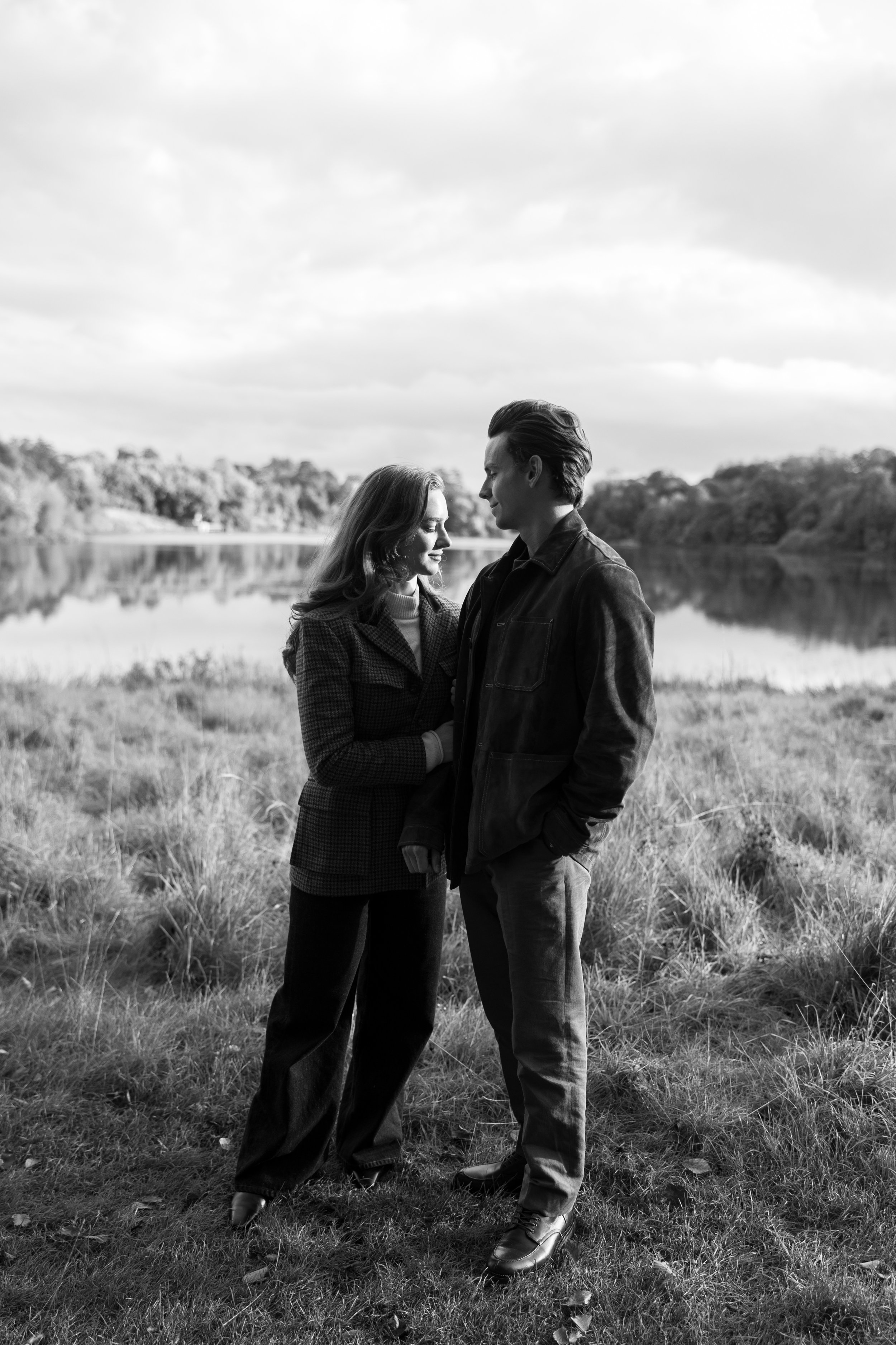 A couple standing close to each other outdoors near a lake with trees in the background, with a cloudy sky overhead.