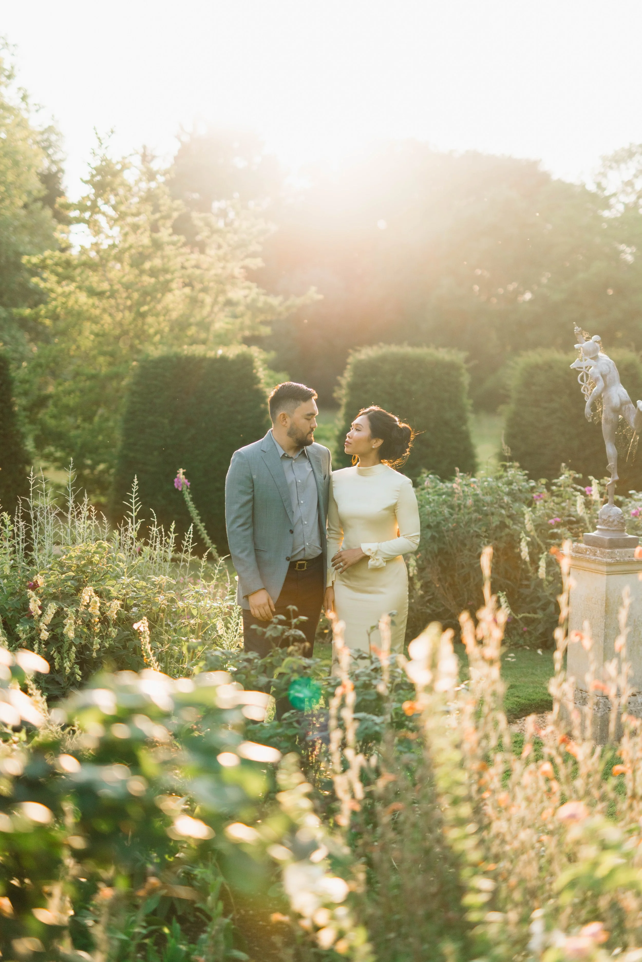 A couple in formal attire standing in a garden during sunset, surrounded by plants and a decorative statue.
