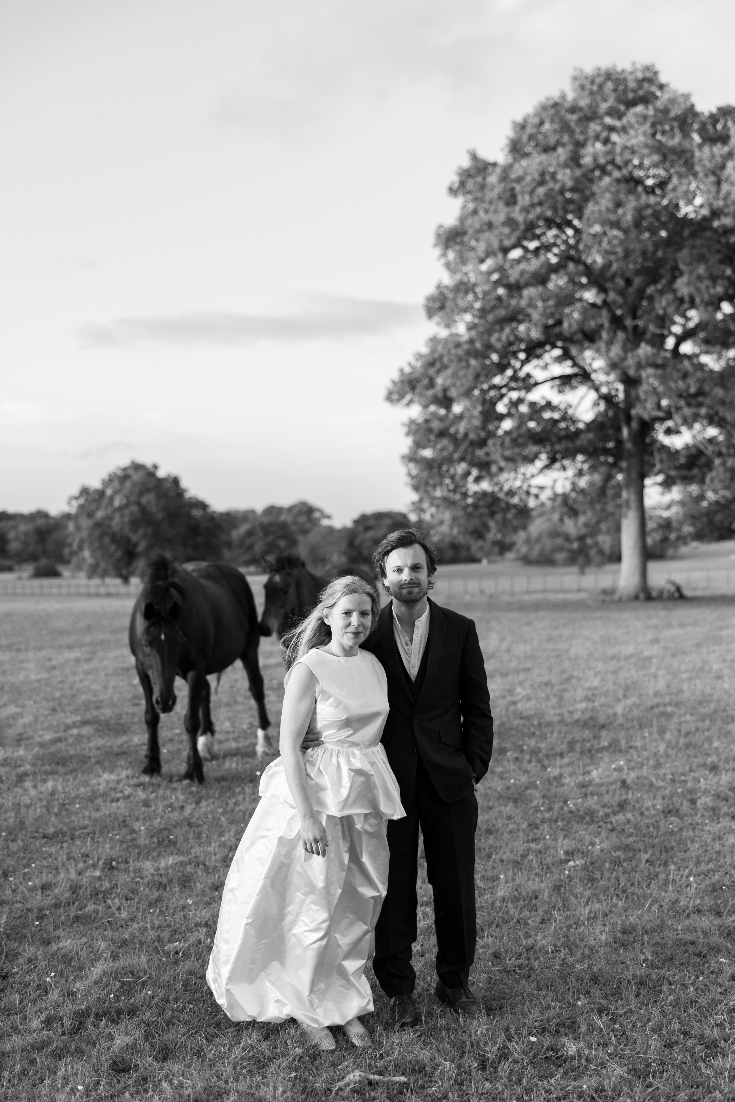 A black and white photo of a woman in a wedding dress and a man in a suit standing in a field with two horses and trees in the background.