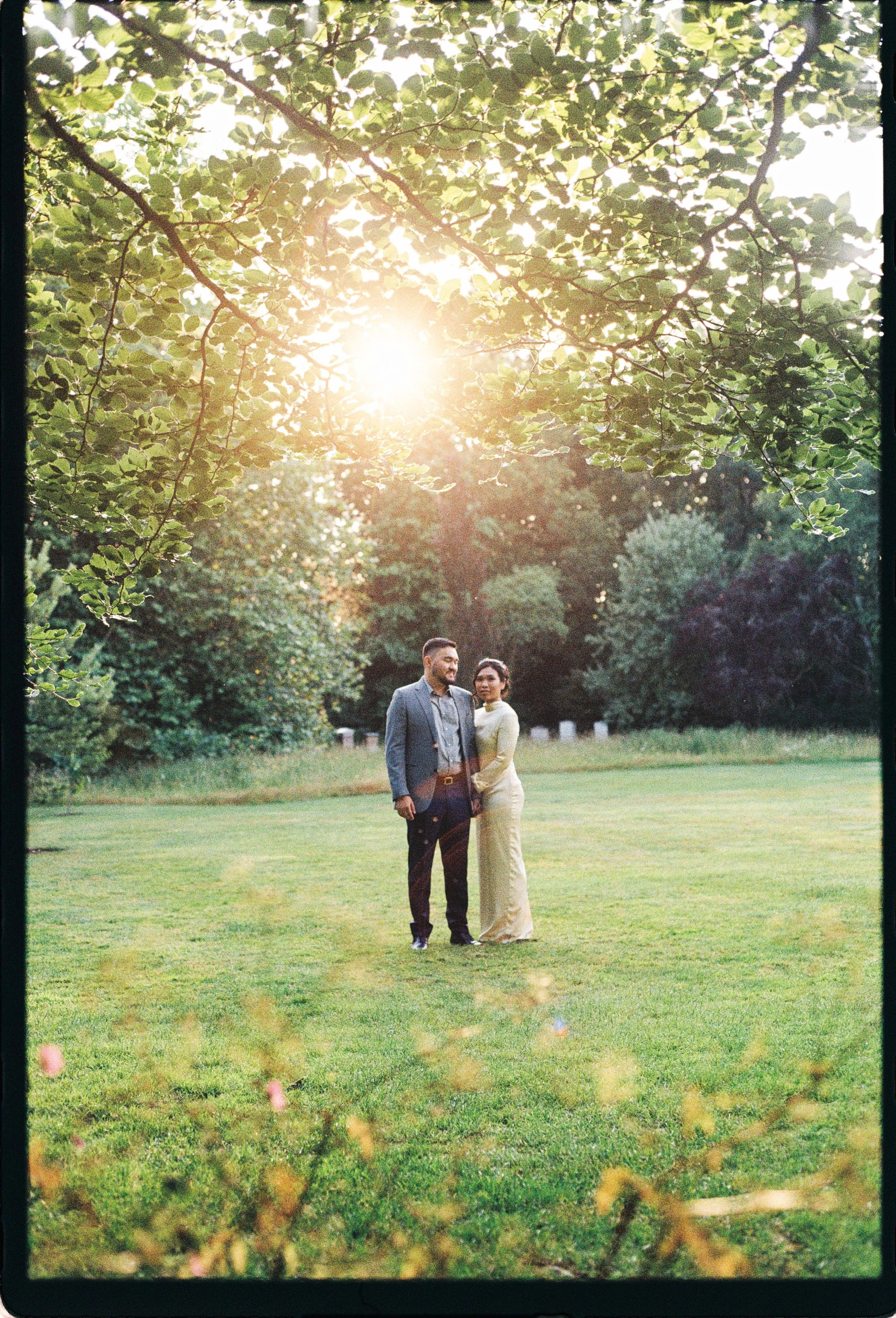 A couple standing hand in hand on a grassy field with trees in the background and sunlight shining through the leaves.