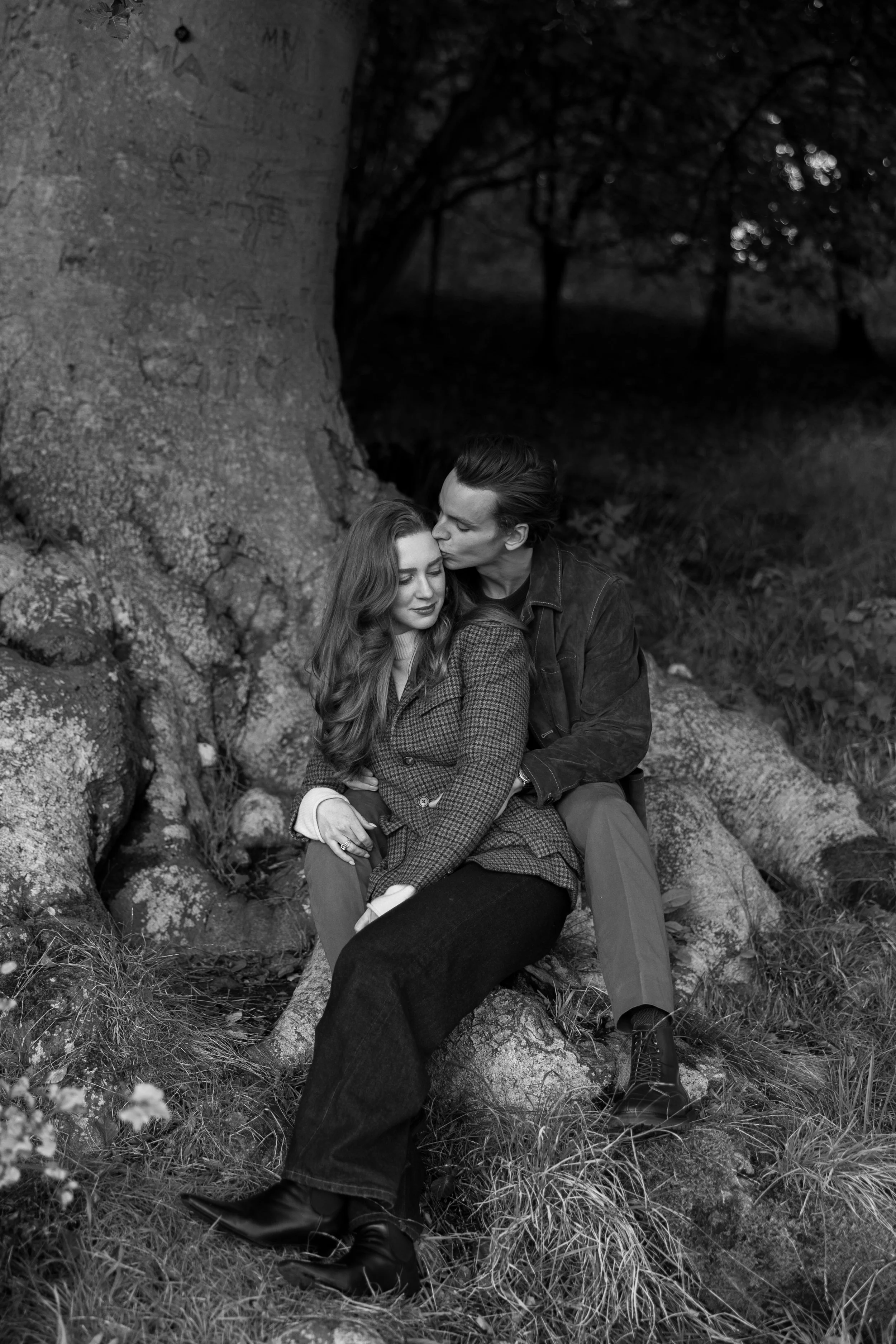 A black and white photo of a man and woman sitting outdoors by a large tree, with the man kissing the woman's forehead. They are surrounded by tall grass and rocks.
