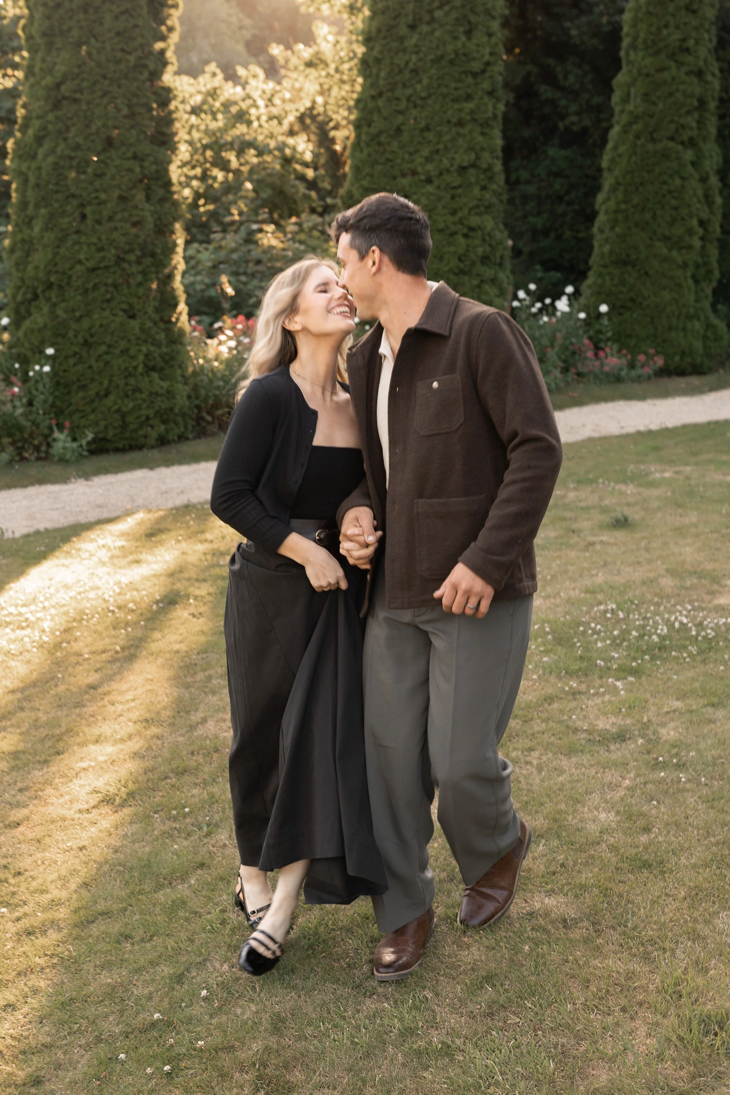 A couple is walking together outdoors, holding hands and smiling at each other, surrounded by trees and flowers during sunset.