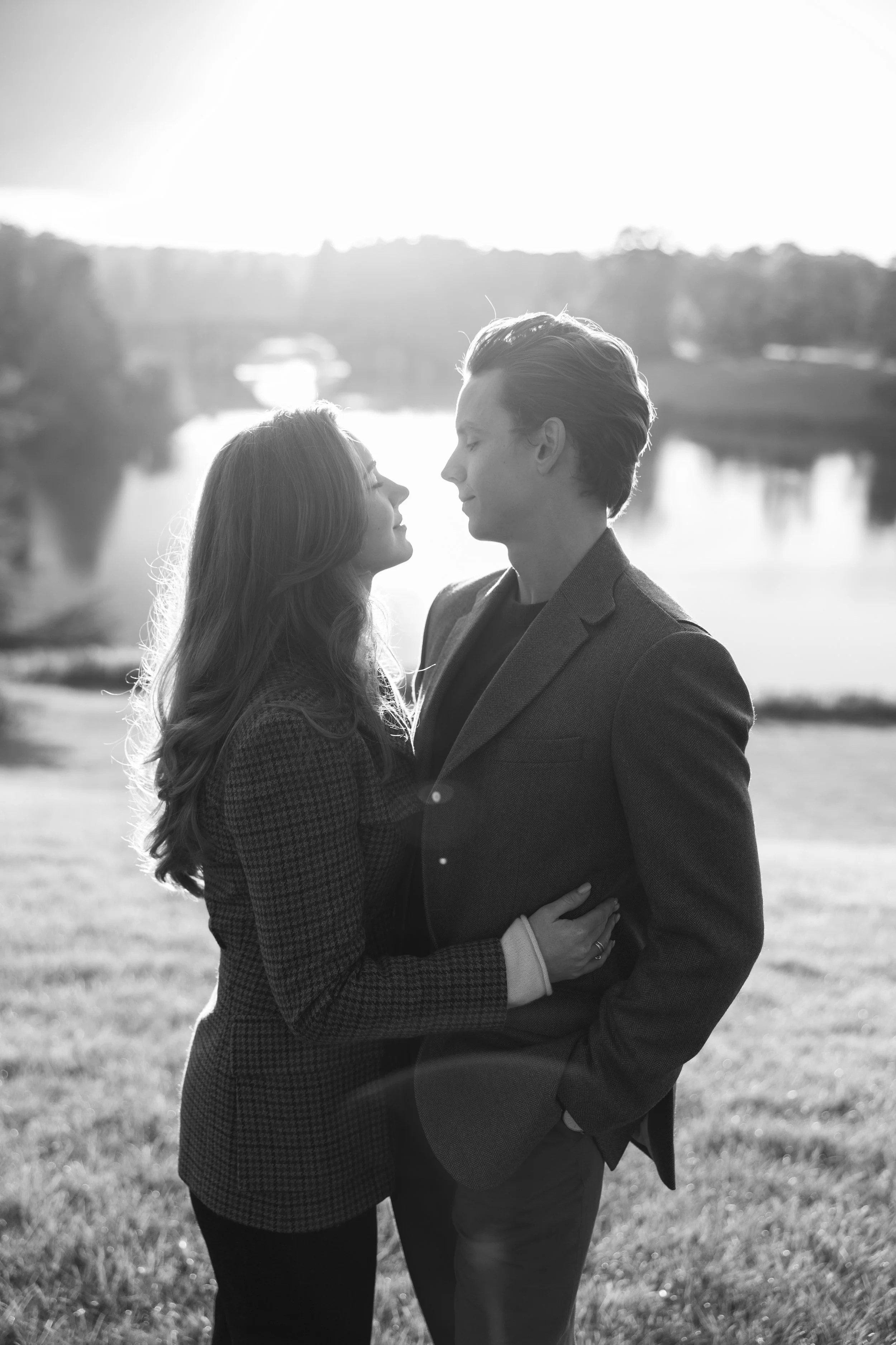 A black and white photo of a man and woman standing close near a lake, with the man gently holding the woman's waist as they look into each other's eyes during sunset.
