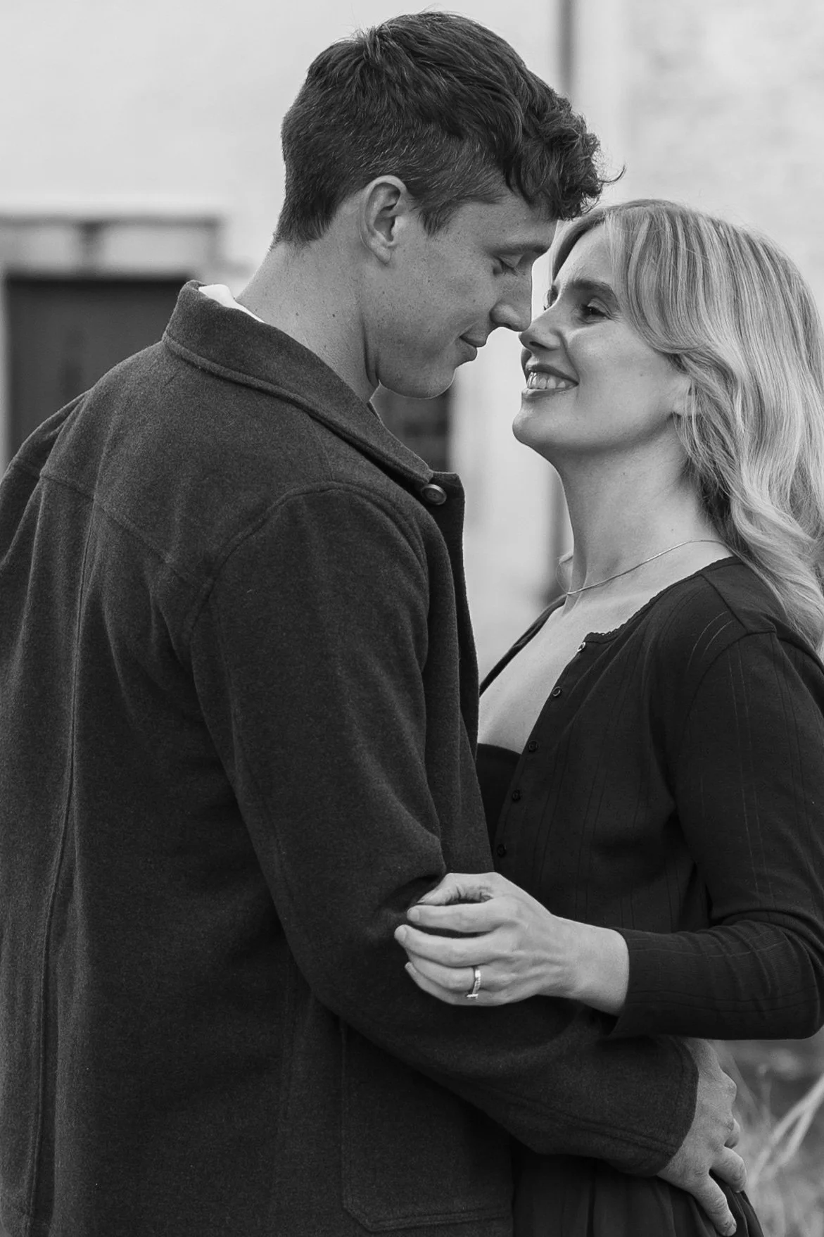 A black and white photo of a young couple standing close together, smiling and about to kiss, with the woman holding the man's arm.