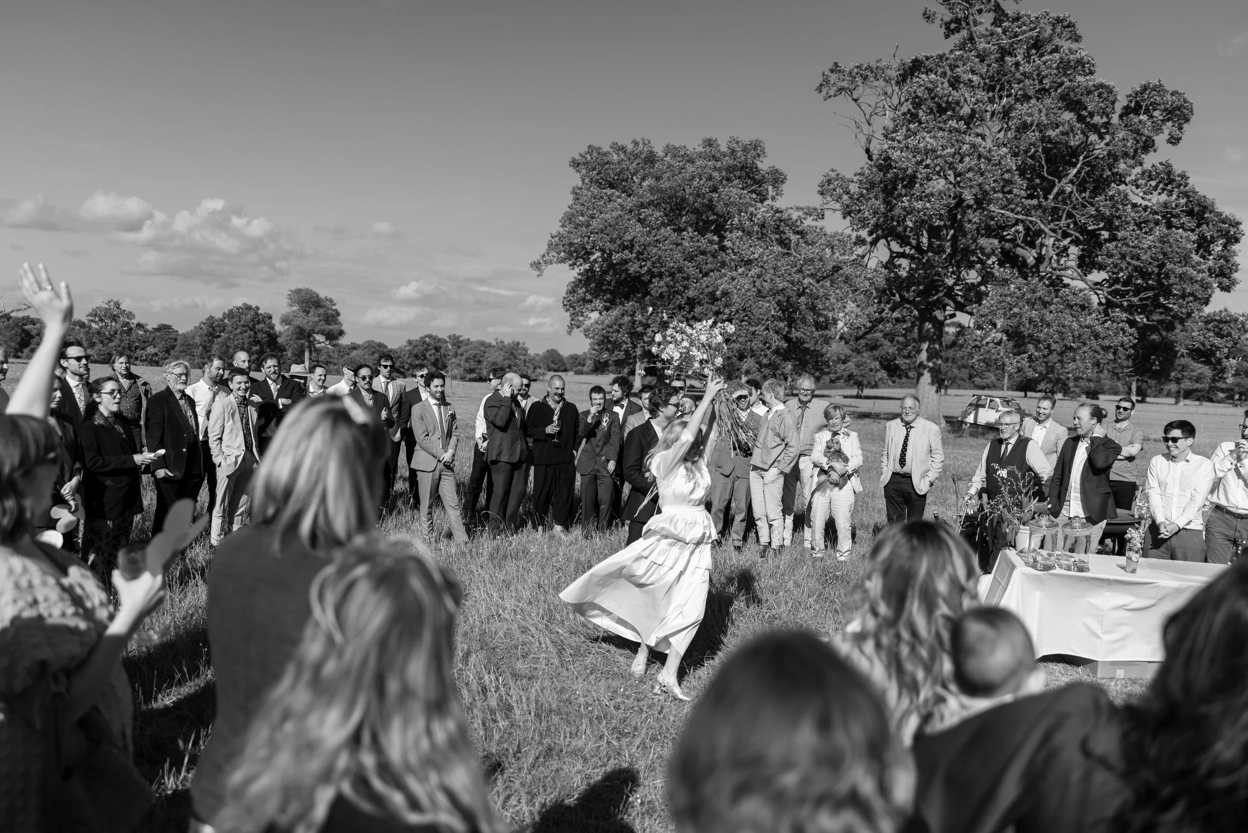 A woman in a white dress dances while holding a bouquet of flowers at an outdoor wedding reception. Guests stand in a semi-circle watching and some are taking photos, with a large tree and open field in the background.