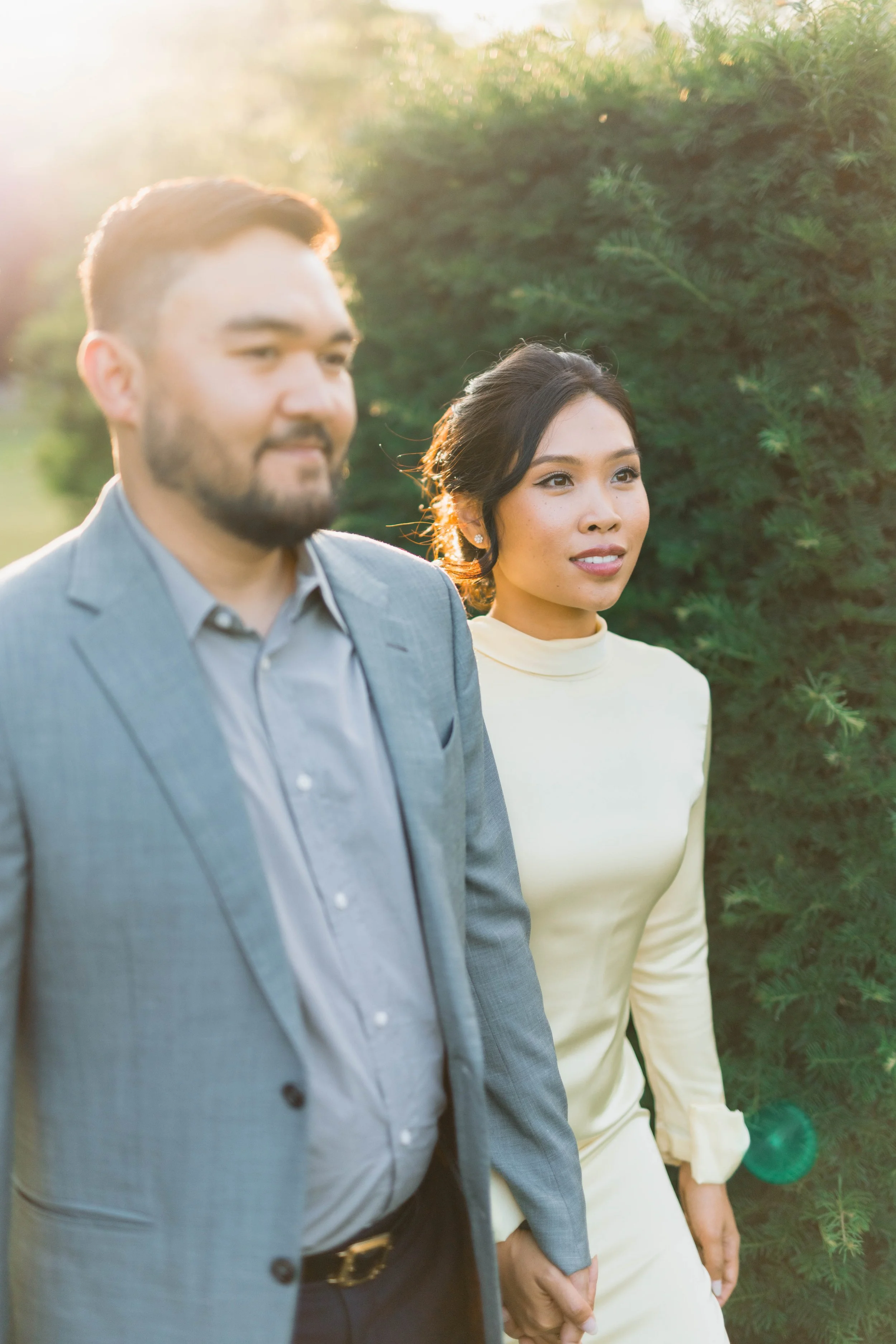 A couple holding hands outdoors during sunset, dressed nicely, with greenery in the background.