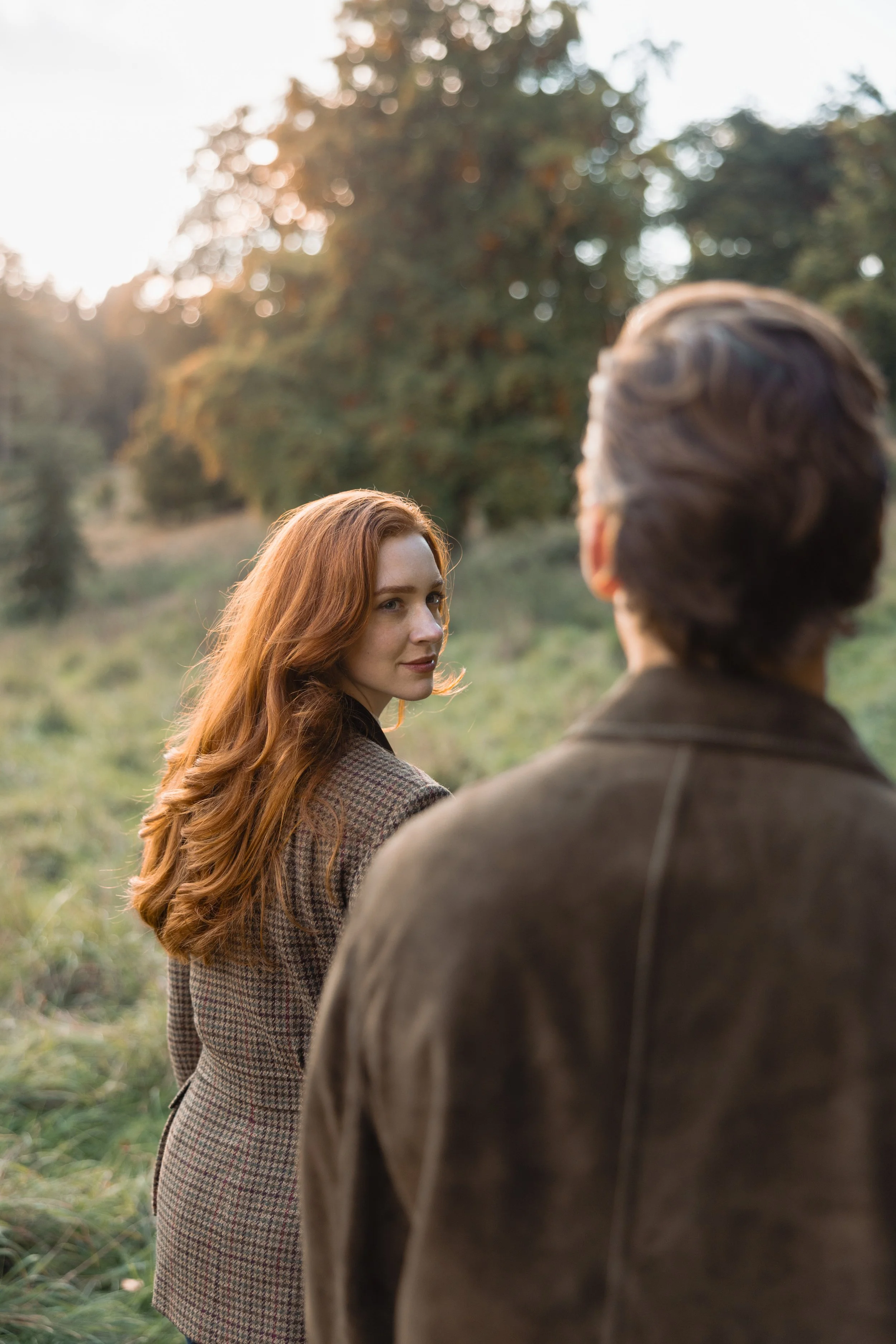 A woman with long red hair and a man with dark hair standing outdoors in a green field with trees in the background, during sunset.