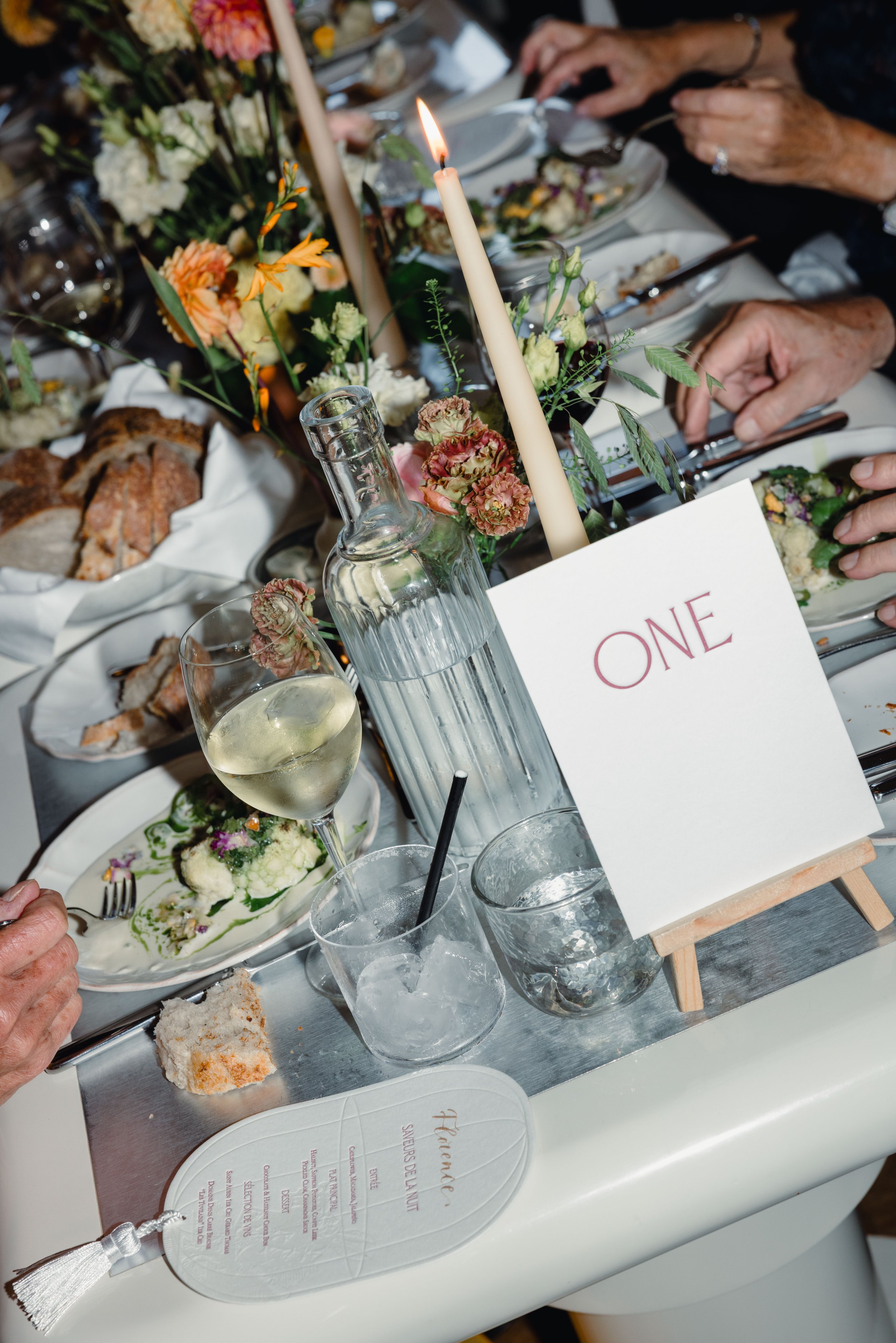 A table setting at a formal event featuring a centerpiece with flowers and tall candle, a glass of white wine, bottles of water, a plate with food, bread, and a table number sign marked 'ONE'.