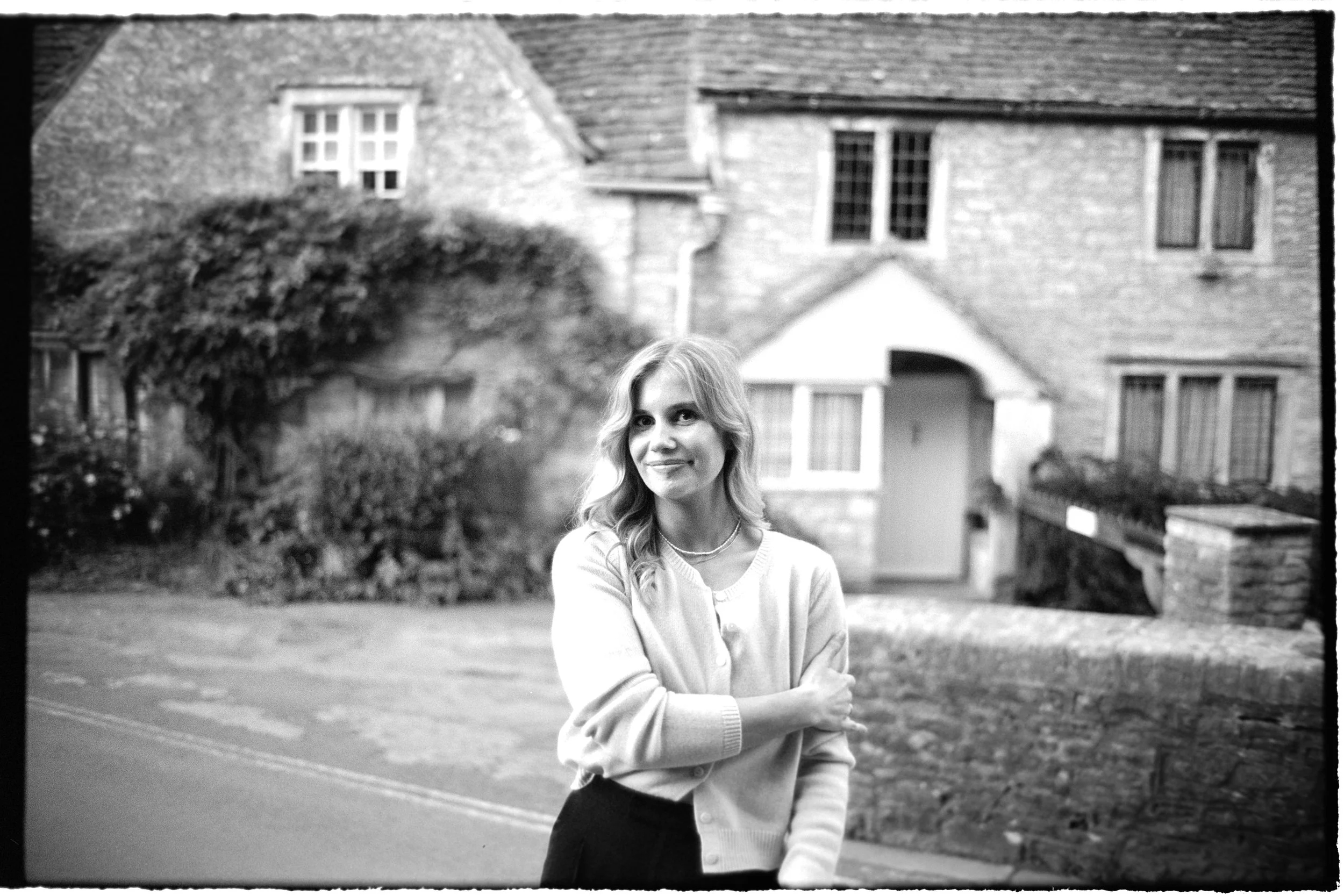 A young woman with wavy hair standing outdoors in front of a stone house with a garden, smiling and crossing her arms.