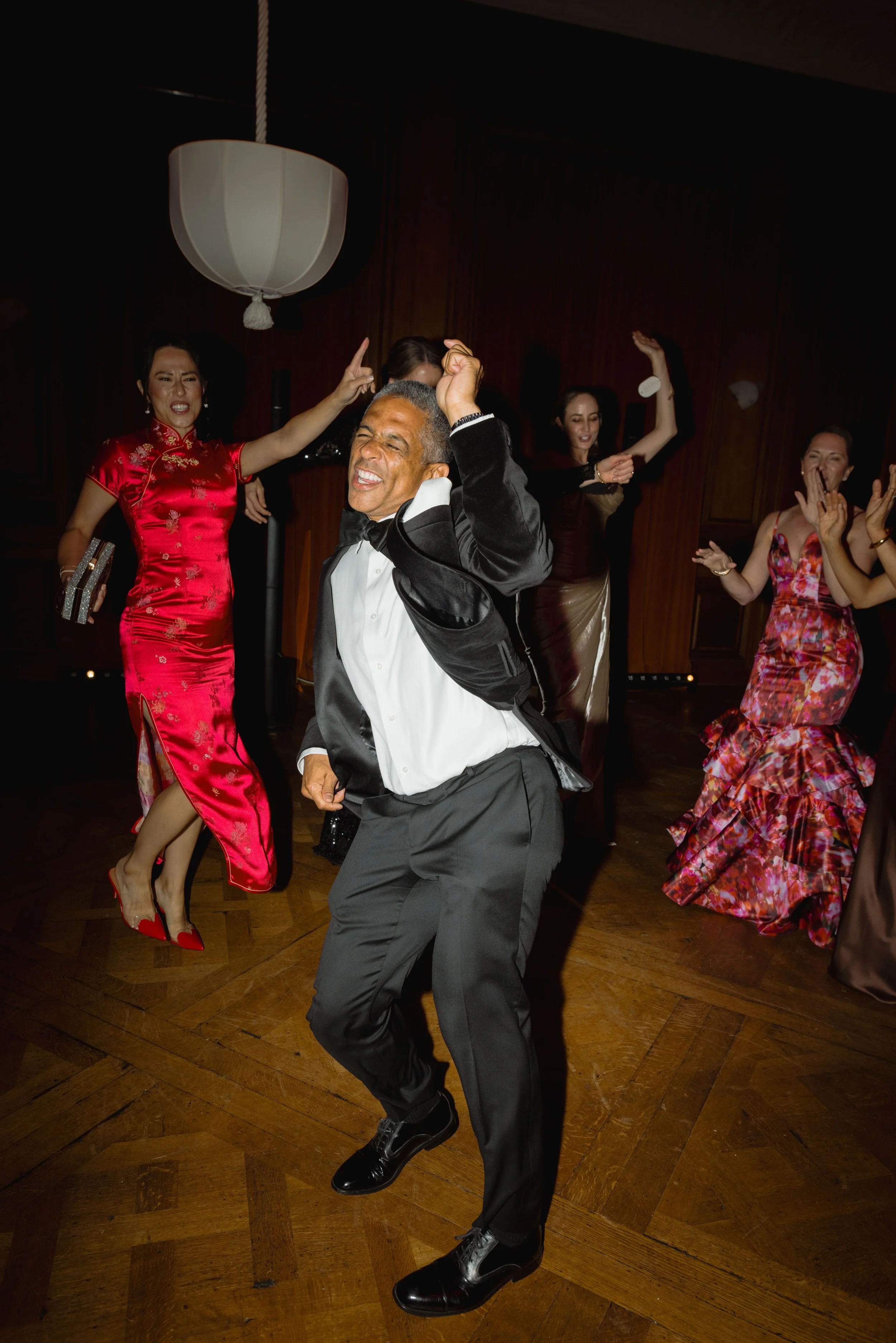 Man dancing joyfully in a tuxedo with women in elegant dresses around him at a party or celebration.