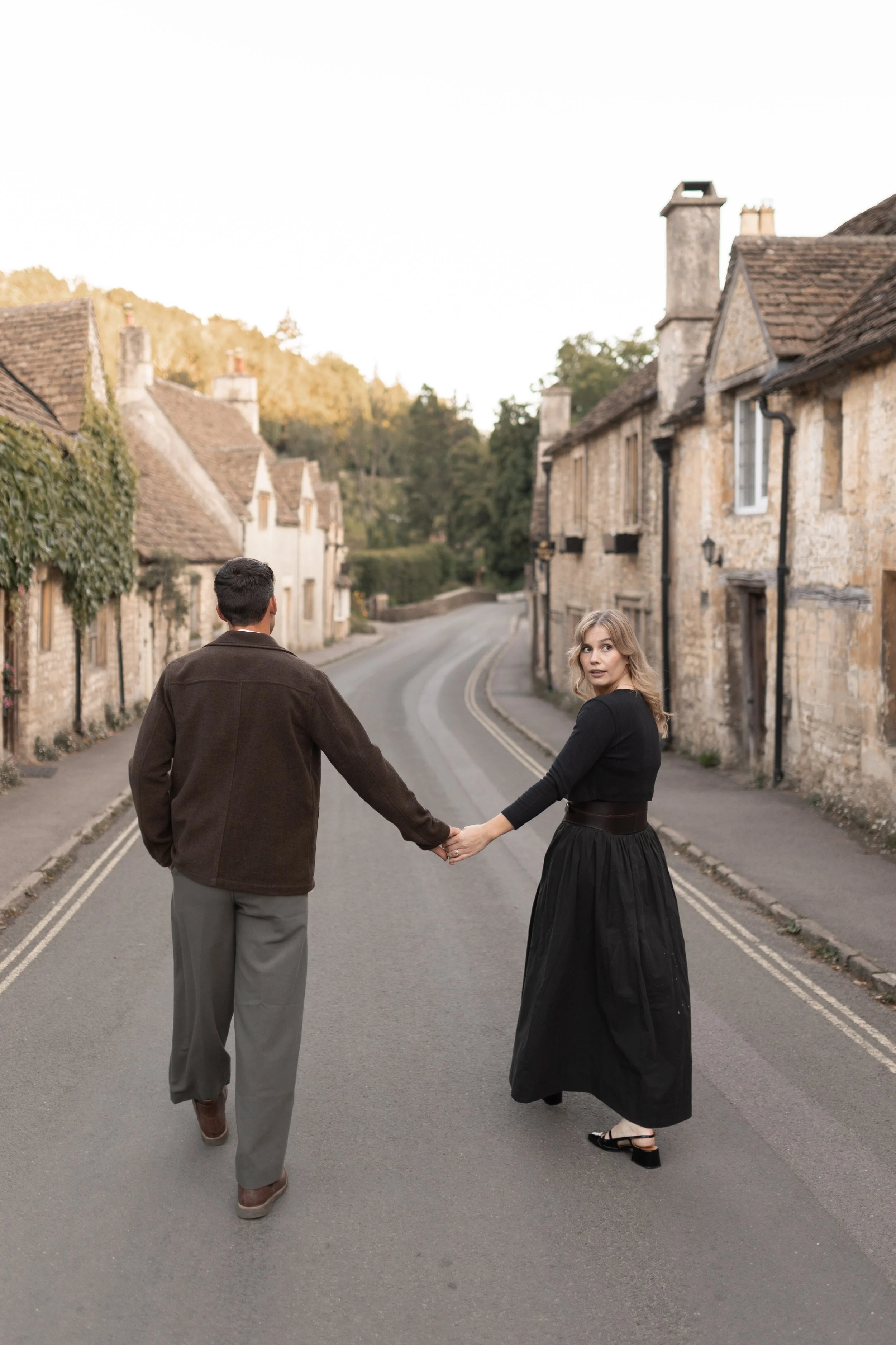 A man and woman holding hands walking across an empty street in a small village with stone houses and greenery, during daylight.