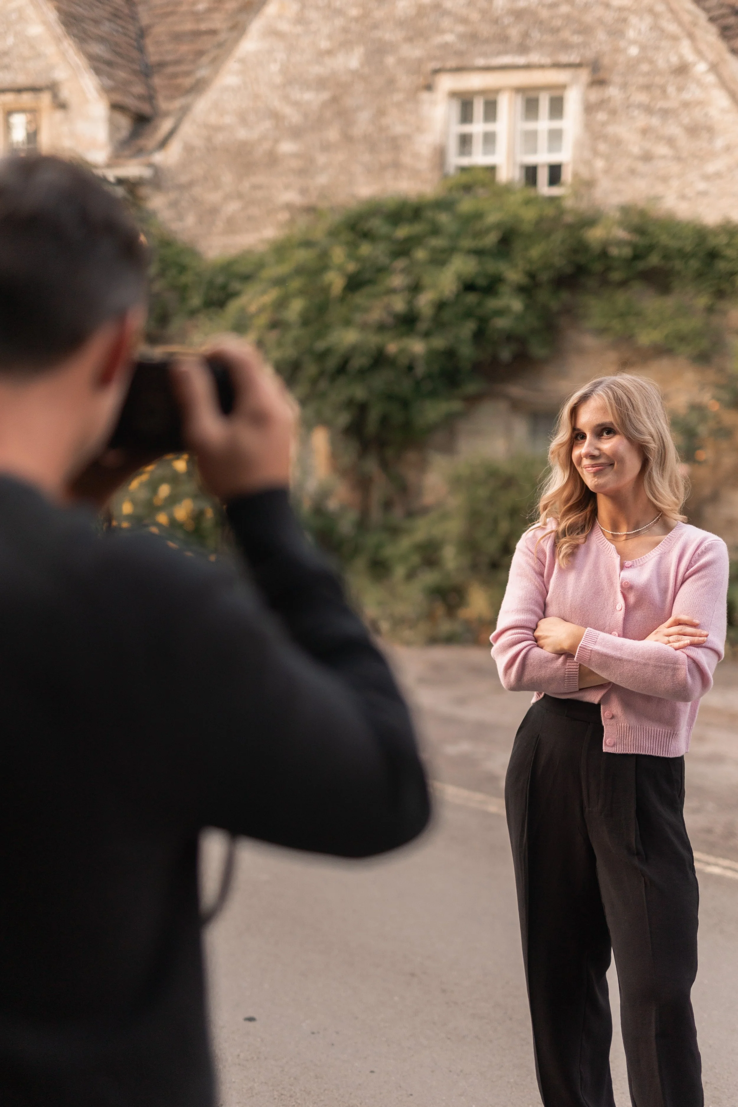 A woman in pink cardigan and black pants posing for a photo outdoors, being taken by a man with a blurred camera in the foreground, with a stone building and greenery in the background.