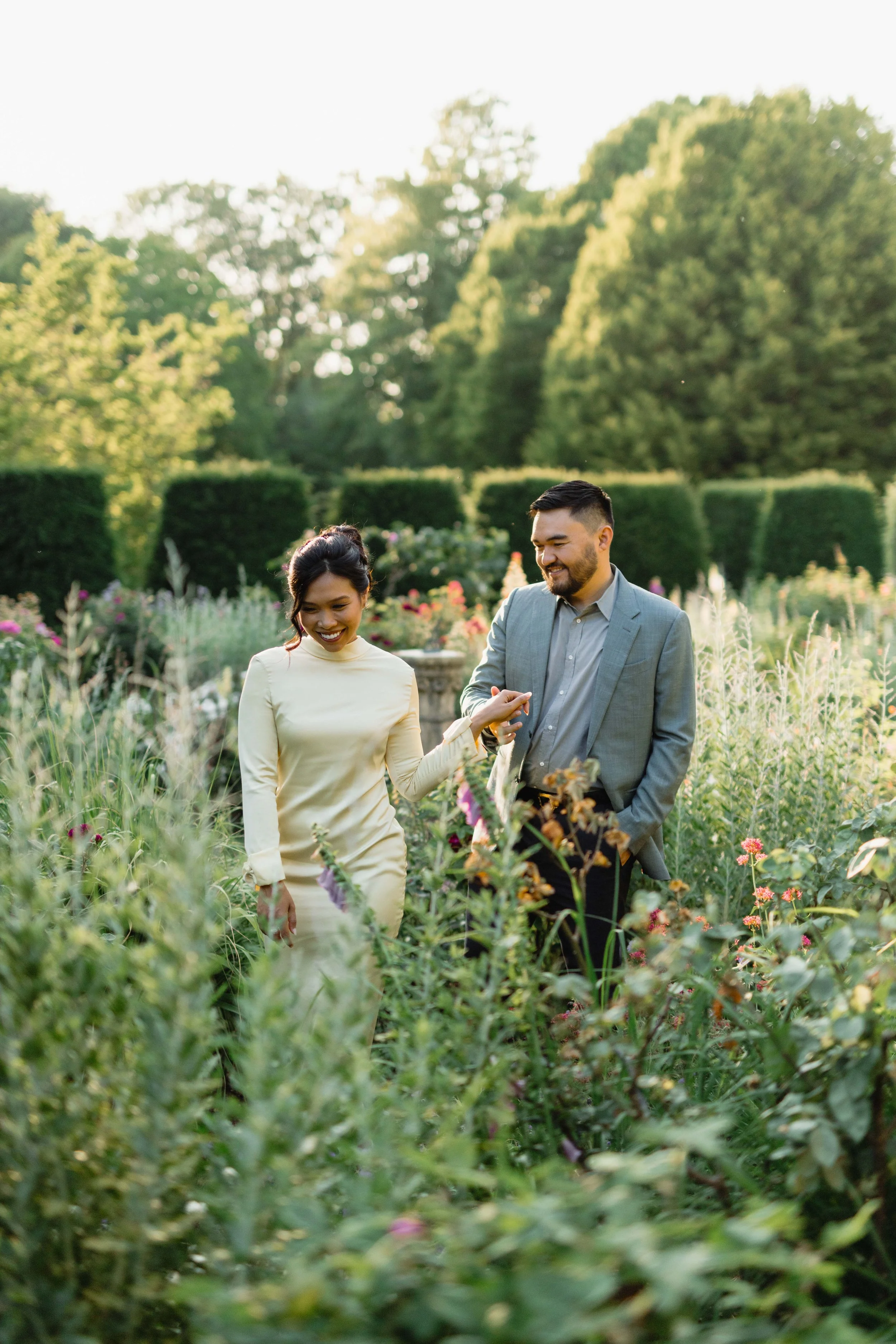 A man and woman walking through a lush garden, smiling and holding hands. The woman is wearing a cream-colored dress, and the man is dressed in a gray suit. The garden is filled with various plants and flowers, with tall trees and greenery in the bac