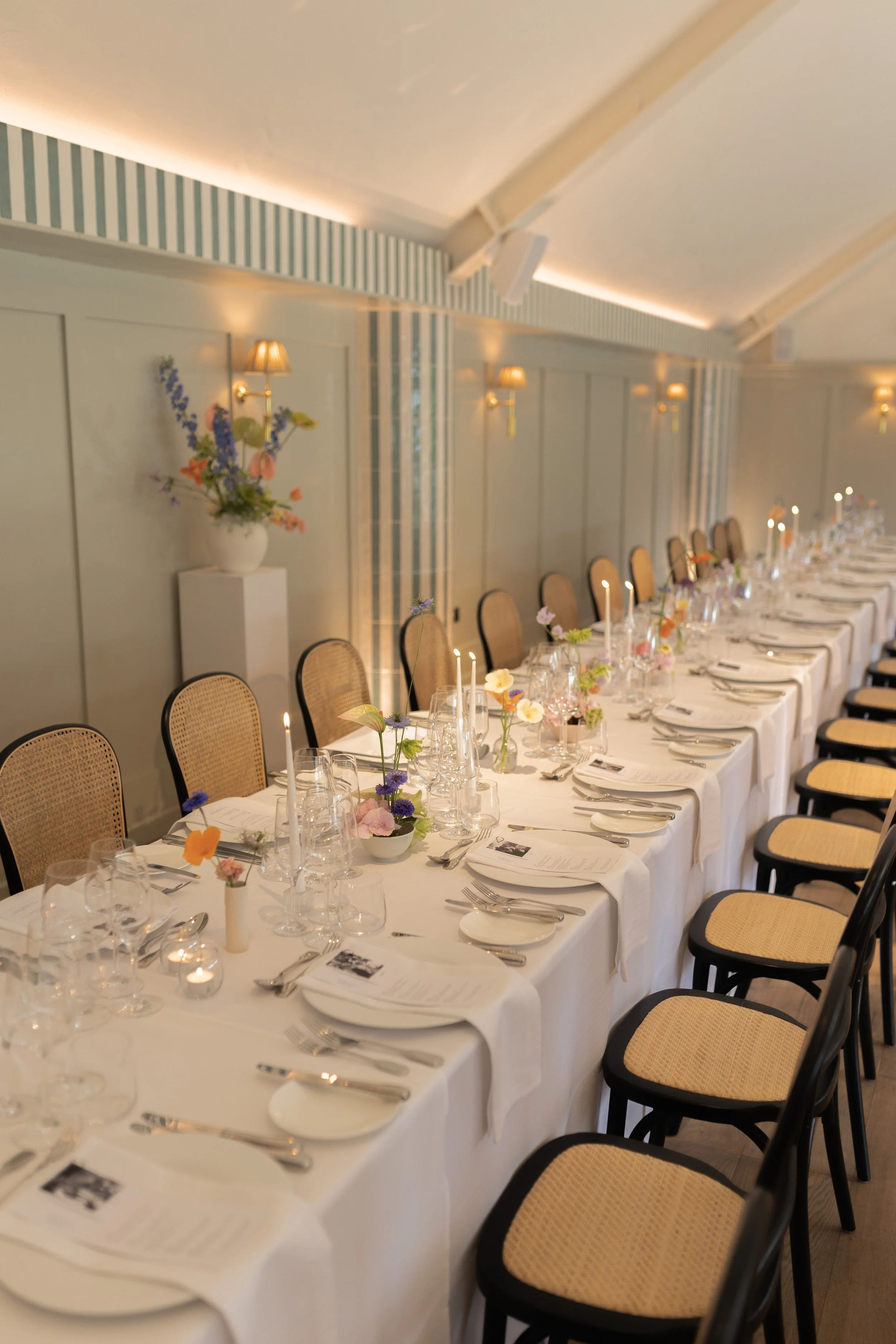 A long formal dining table set for a special occasion with white tablecloths, flower centerpieces, candles, and neatly arranged plates, silverware, and glassware, in a room with soft lighting and pastel-colored walls.