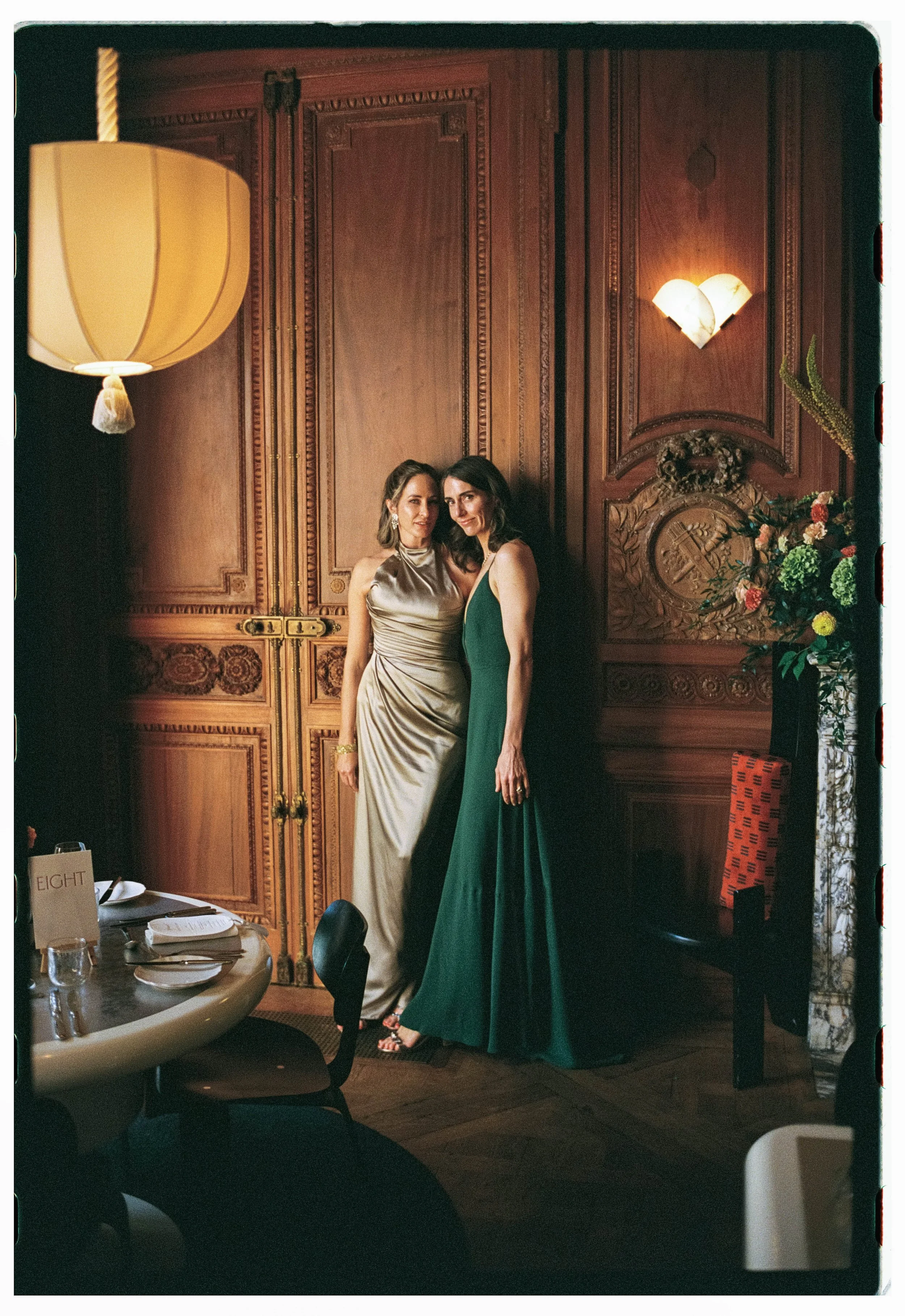 Two women in elegant dresses standing close together in a richly decorated wooden-paneled room with a large floral arrangement on the right, soft lighting, and a round dining table with place settings in the foreground.