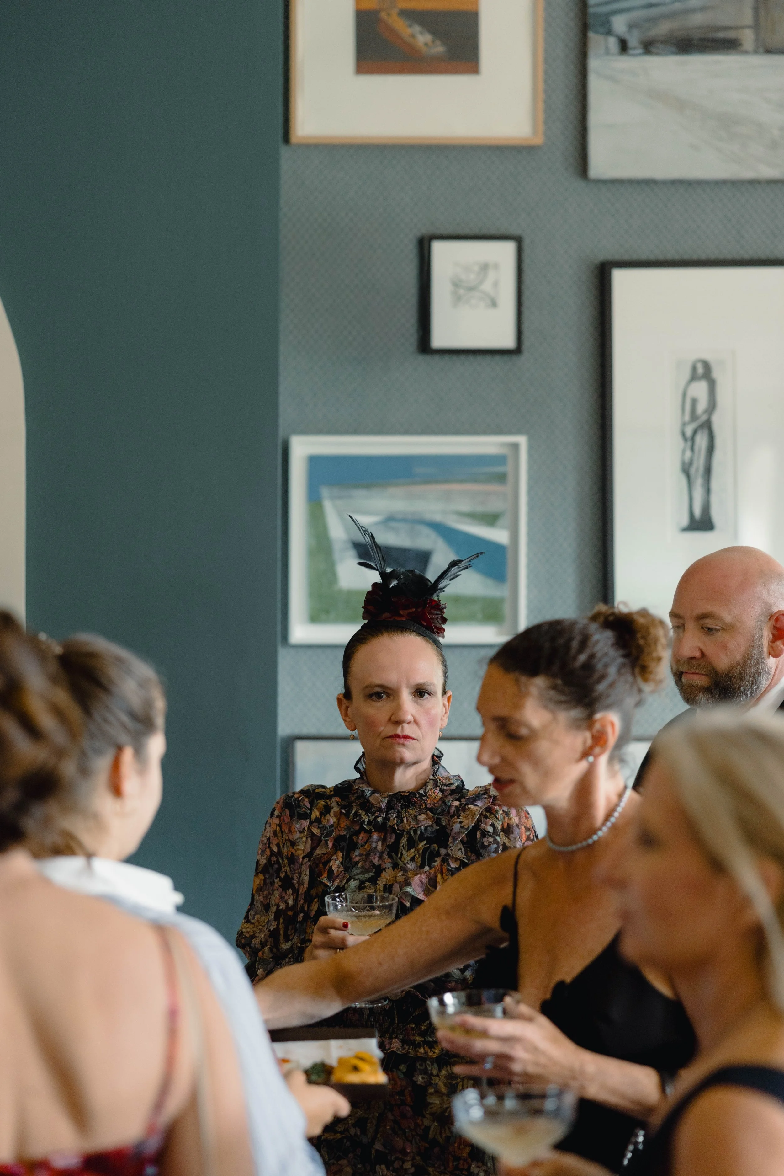 A group of people at a wedding, with women in elegant attire holding cocktails, in a decorated indoor setting with framed artwork on the wall.