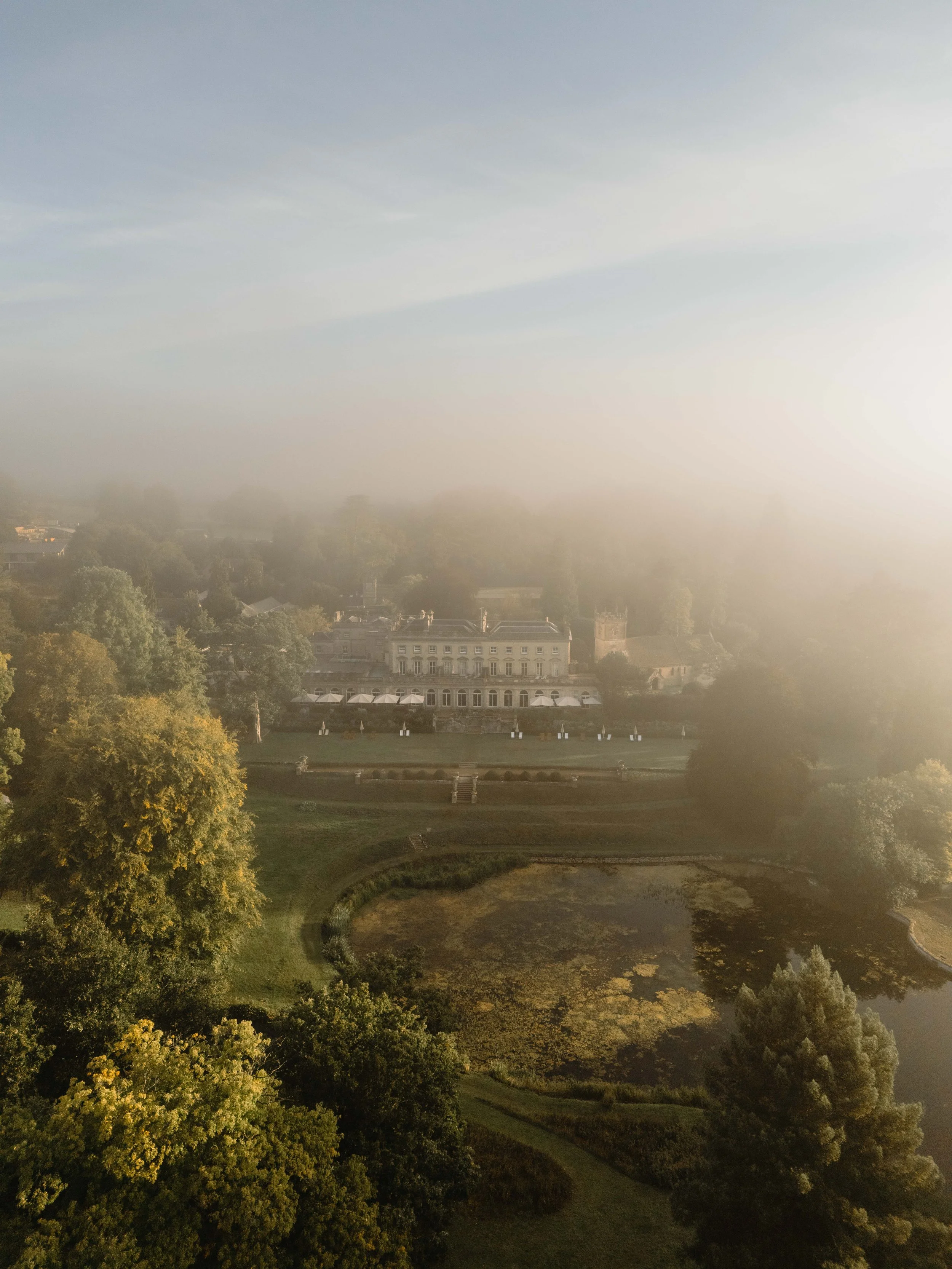 Aerial view of Cowley Manor Experimental a large estate with a mansion, surrounded by trees, a pond, and a hazy sky.