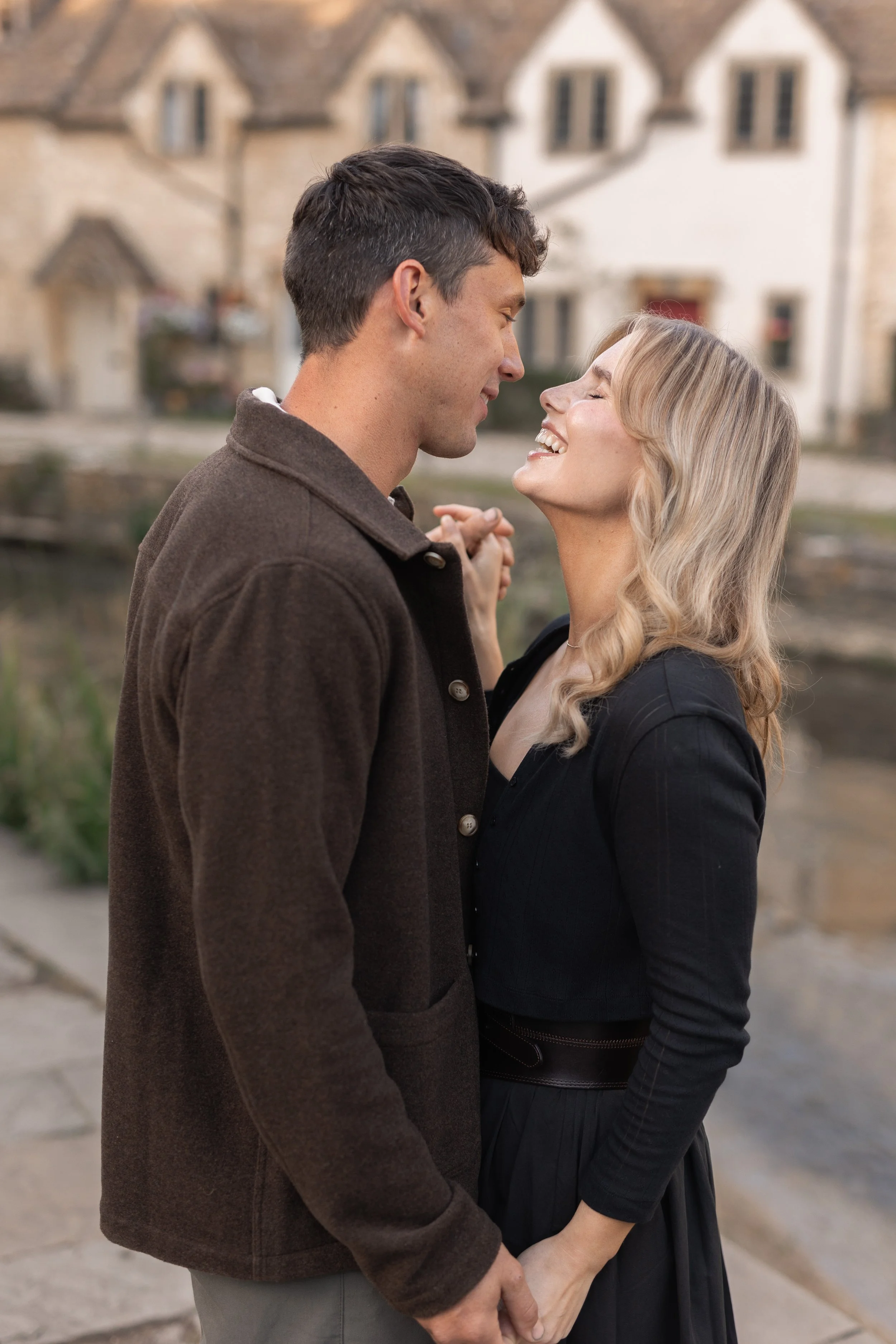 A couple standing closely together outdoors, holding hands, smiling and gazing at each other, with houses in the background.