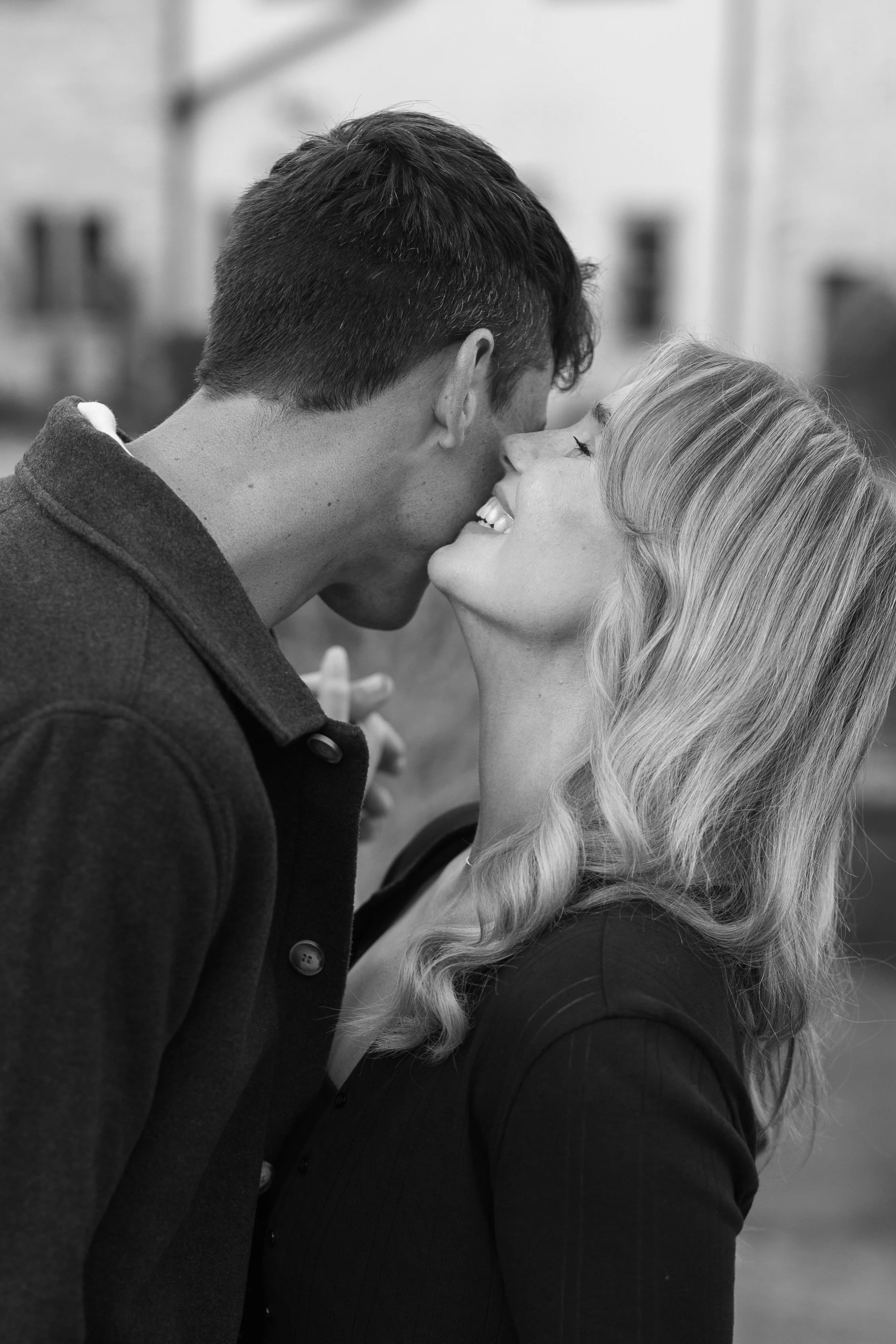 A black and white photo of a man and woman about to kiss outdoors, with the woman smiling and the man leaning in.