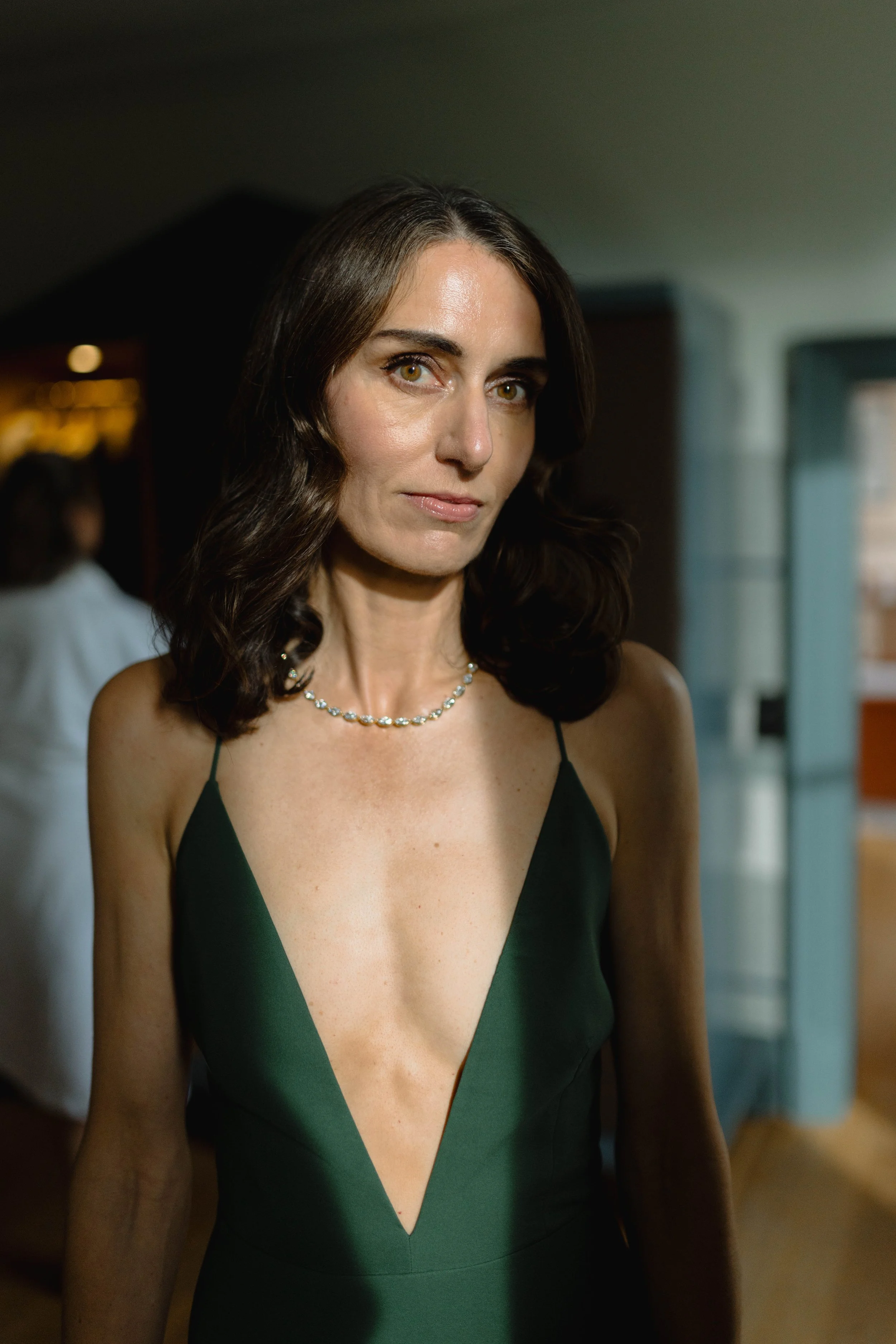 A woman with wavy brown hair wearing a dark green dress with a deep V neckline and a pearl necklace, standing indoors with a neutral expression.