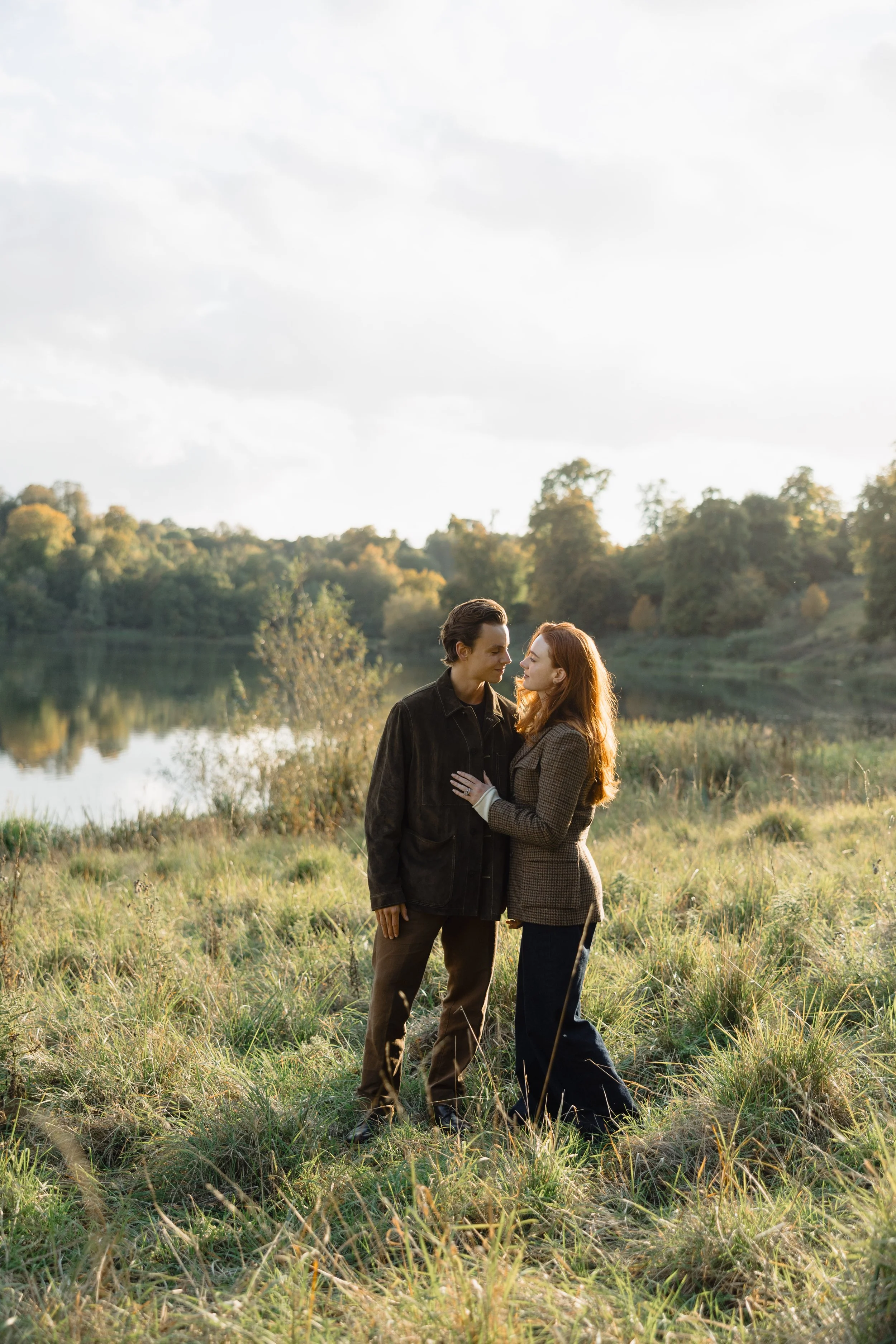 A couple in vintage clothing standing close by a lake in a grassy field, gazing at each other during sunset.
