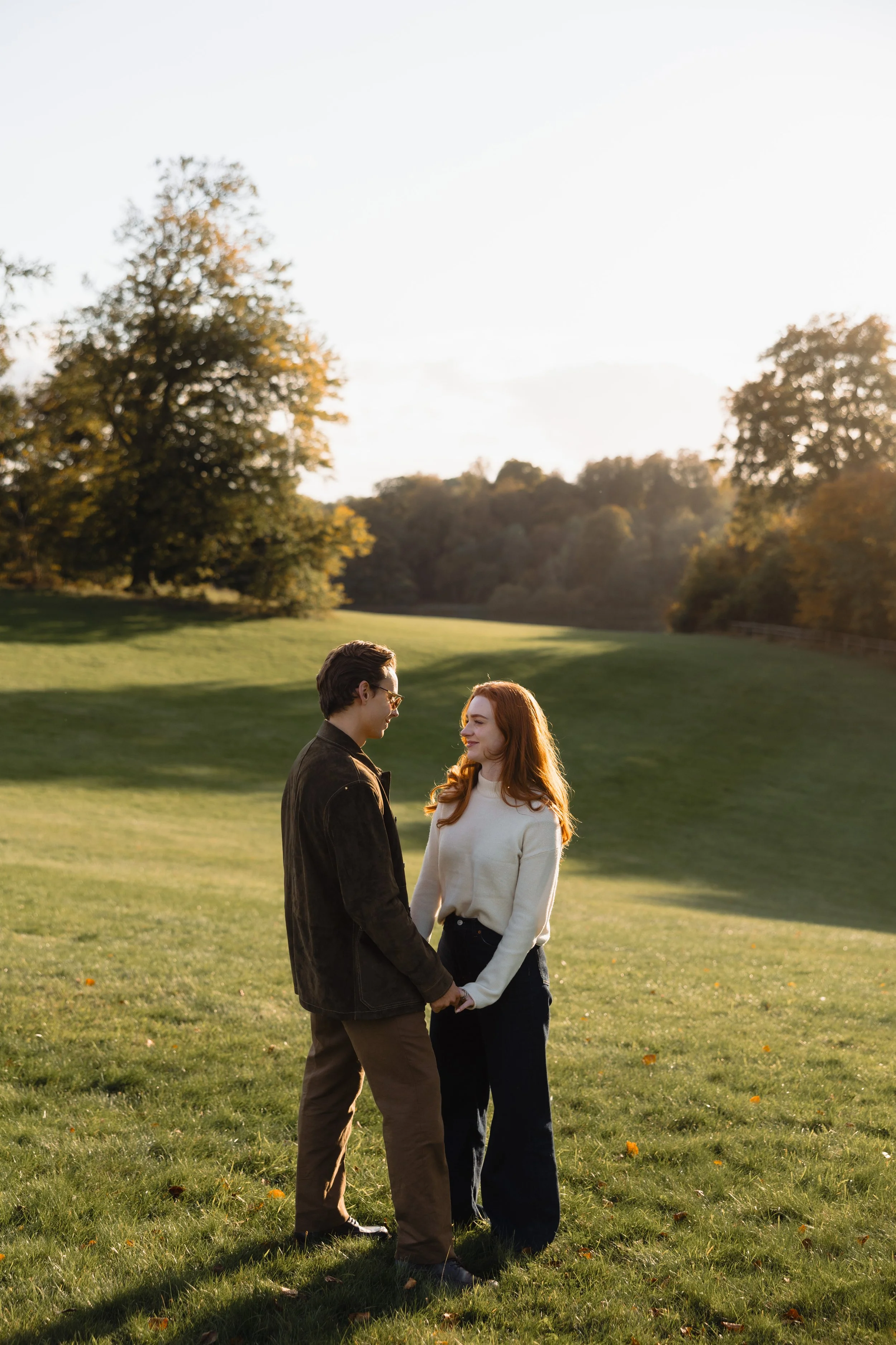 A couple holding hands and facing each other in a grassy park during sunset, with trees in the background.