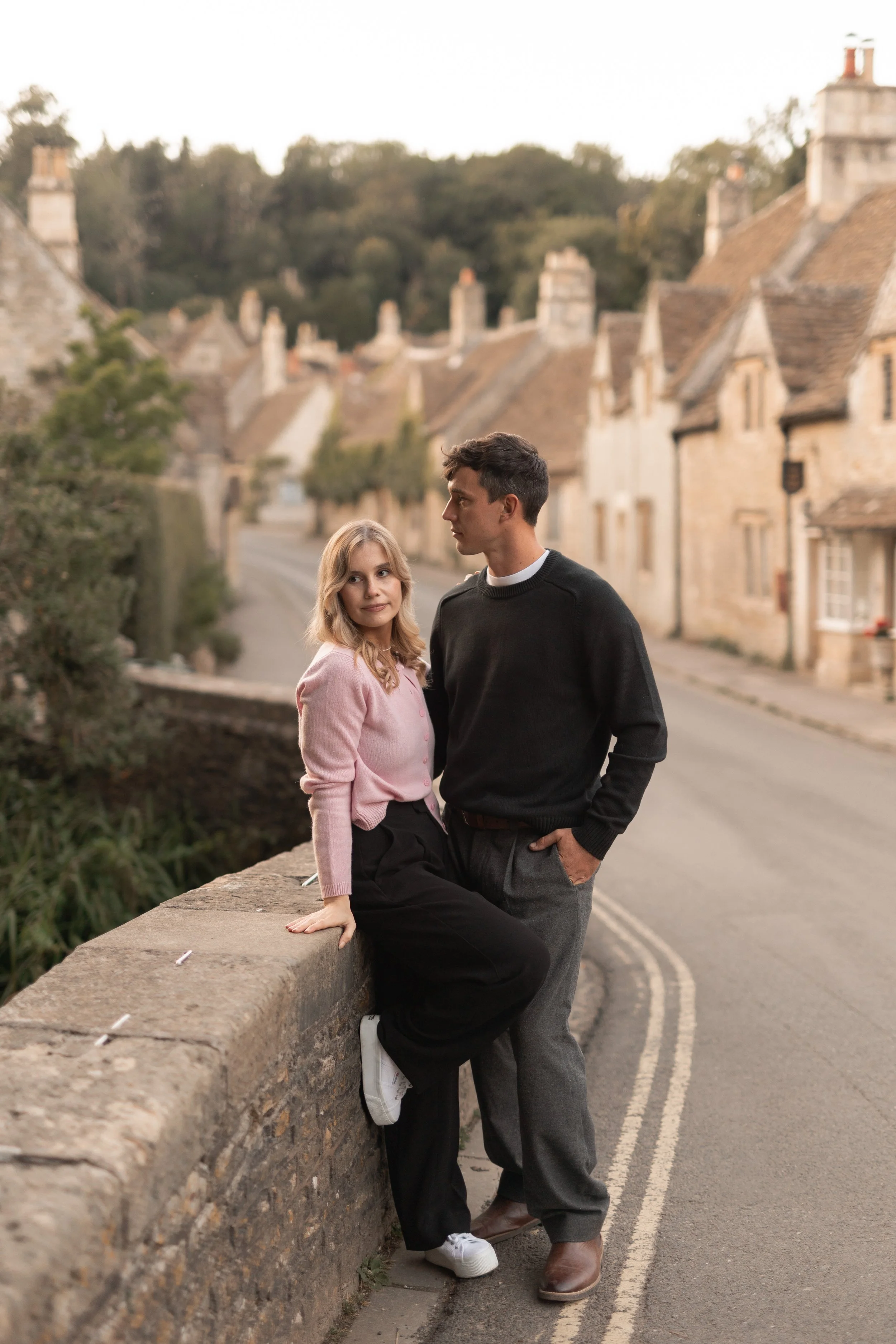 A young woman perched on a stone wall and a young man standing next to her on a street in a quaint village with stone houses and a hilly landscape in the background.