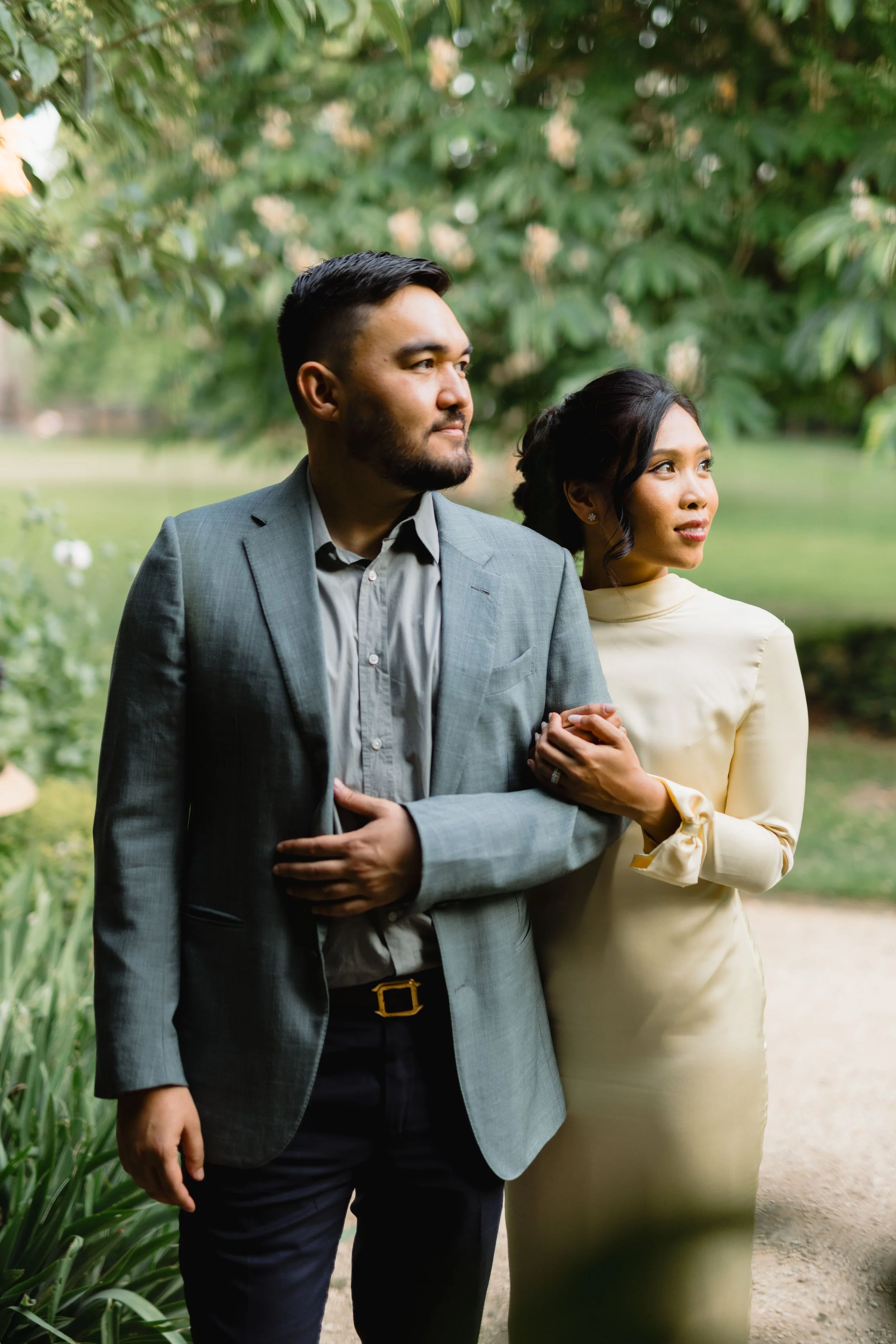A man and woman in formal attire standing outdoors in a park-like setting, with trees and a pond in the background, holding hands and looking in different directions.