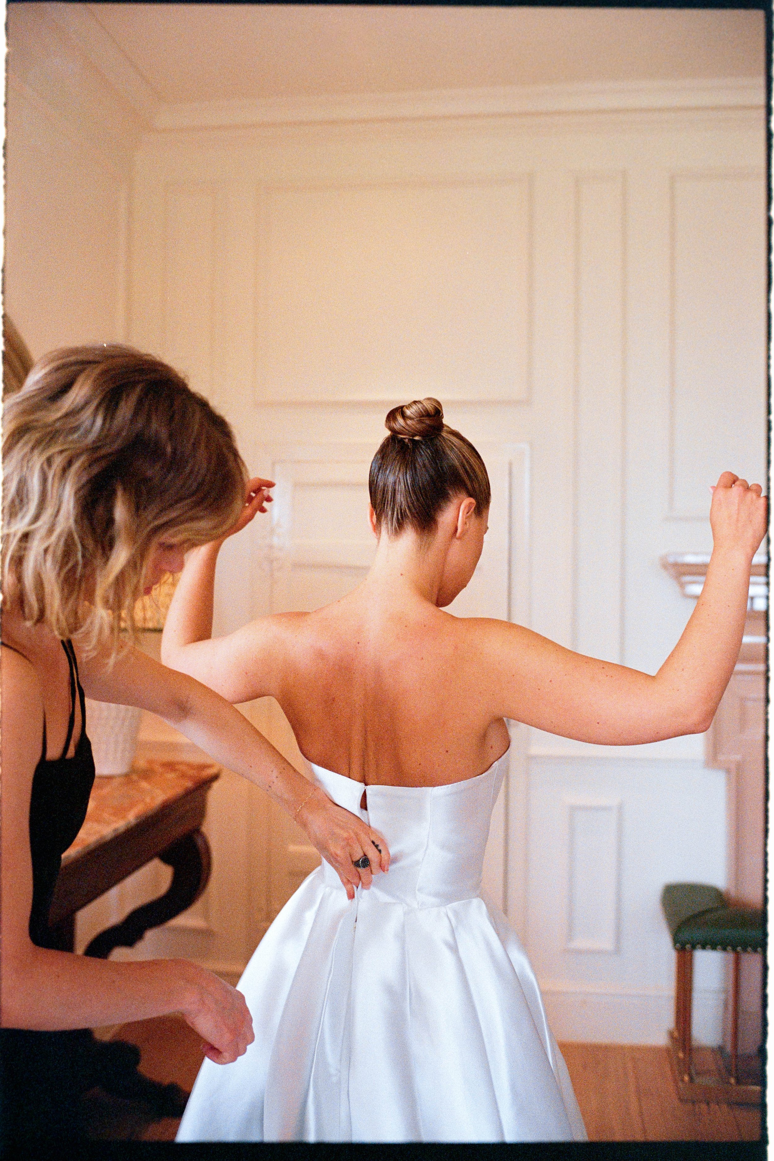 A bride in a strapless white wedding dress helping her get dressed, assisted by a woman in a black dress, in a cozy room with white paneled walls and a small green chair.