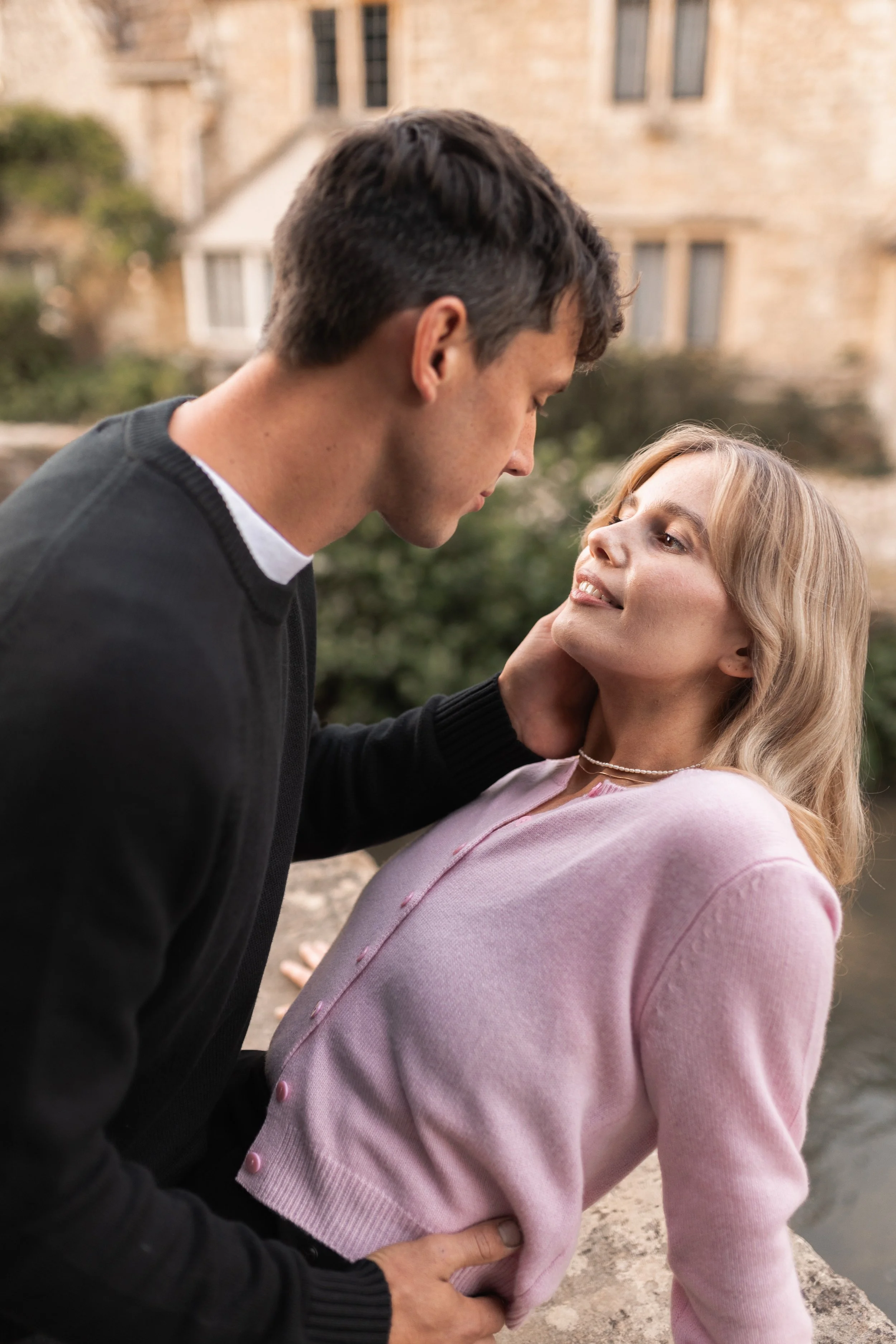 A man and woman face each other closely outdoors with a building and greenery in the background.