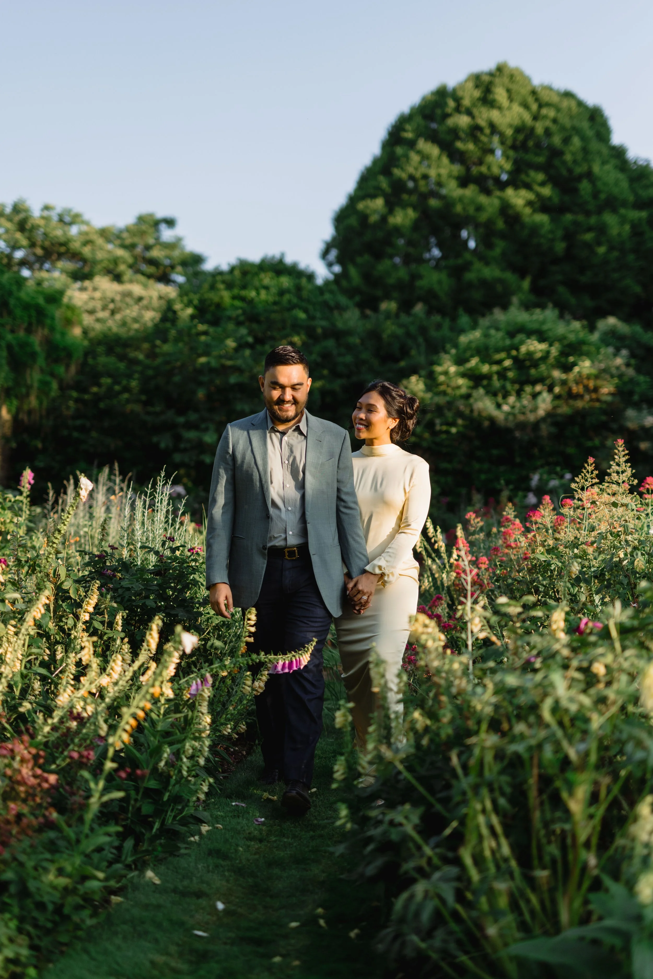A smiling couple holding hands walking through a flower garden with green trees in the background.