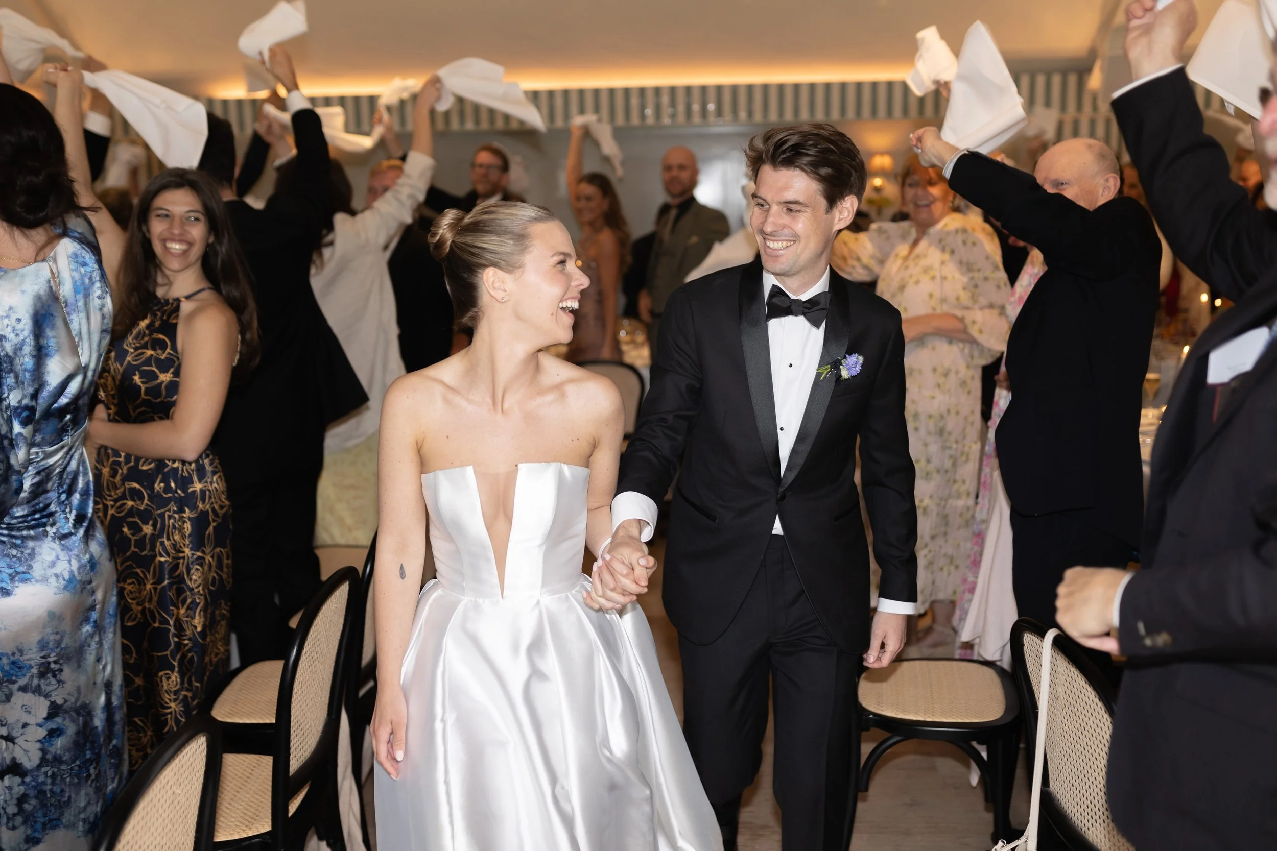 A newlywed couple holding hands and smiling at each other while walking through a celebration with guests raising napkins in the air.