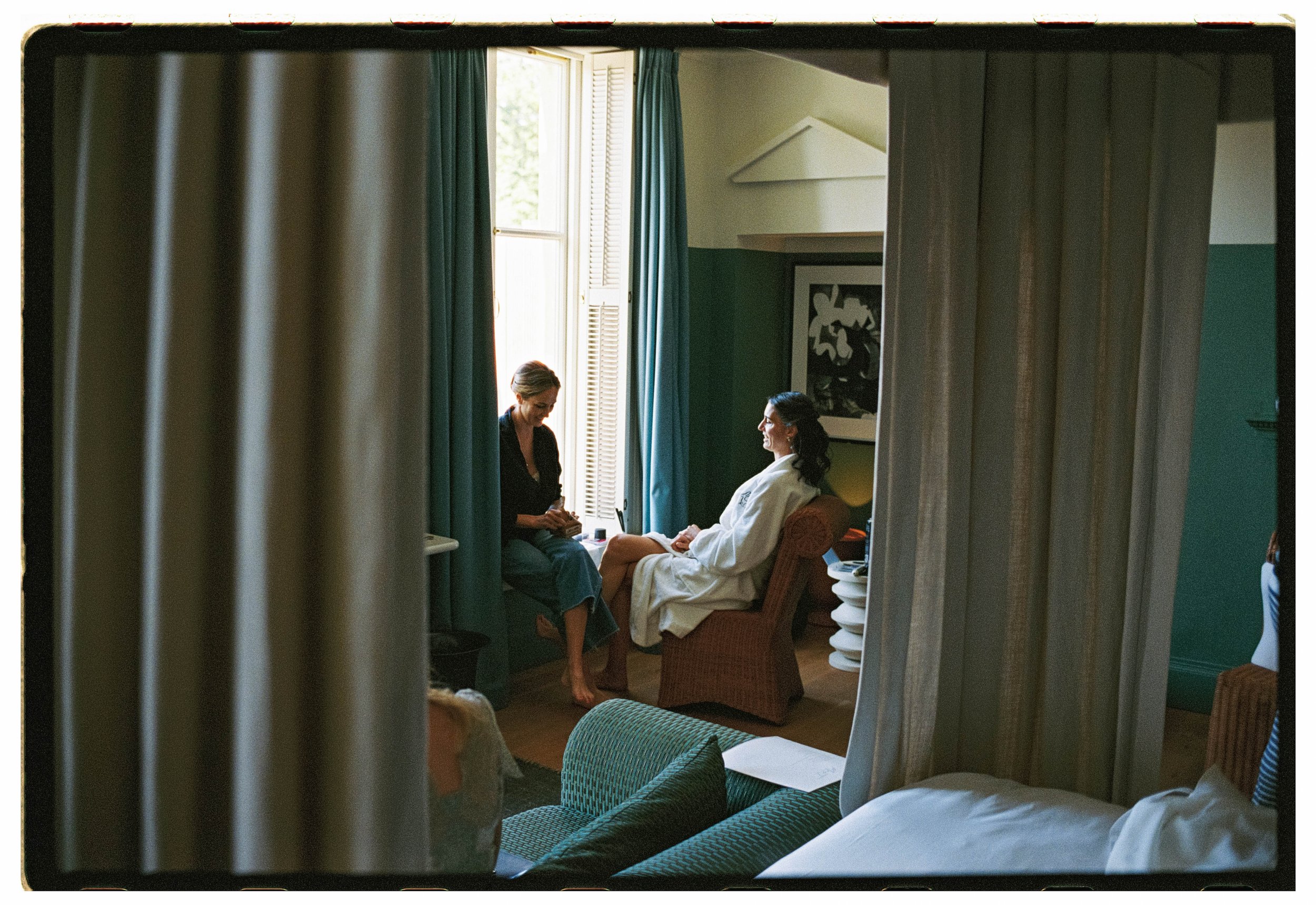 Two women in a hotel room, one sitting on a chair wearing a white bathrobe and the other sitting on the windowsill wearing casual clothes, engaging in conversation. The room has large windows with open shutters, teal curtains, and framed artwork on t
