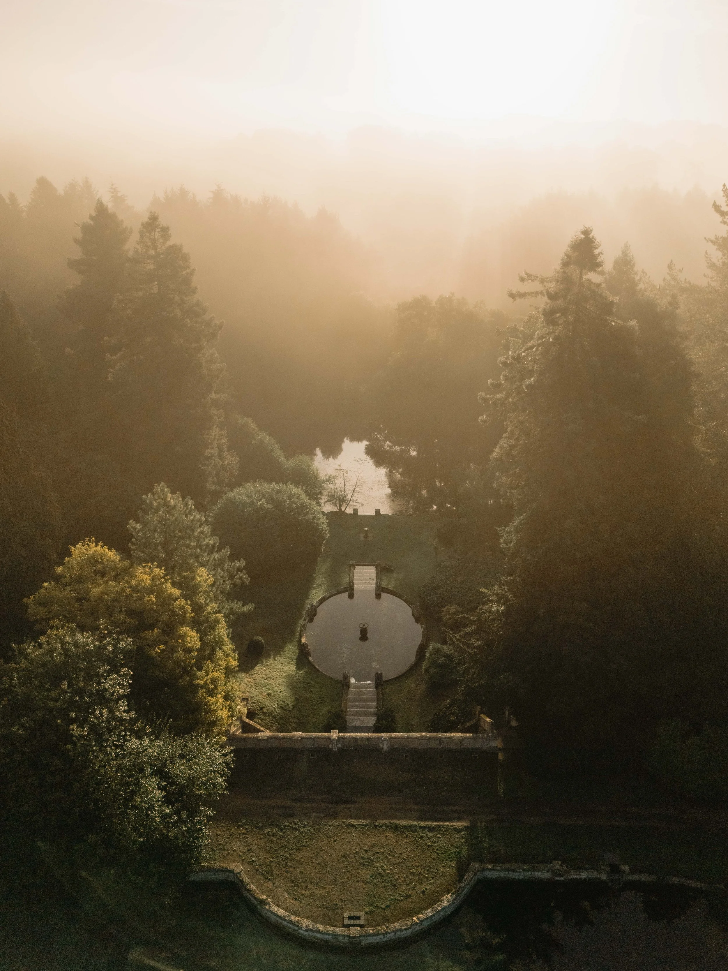 Aerial view of the park at Cowley Manor Experimental
 with two circular ponds connected by a small bridge, surrounded by lush trees and foggy weather.