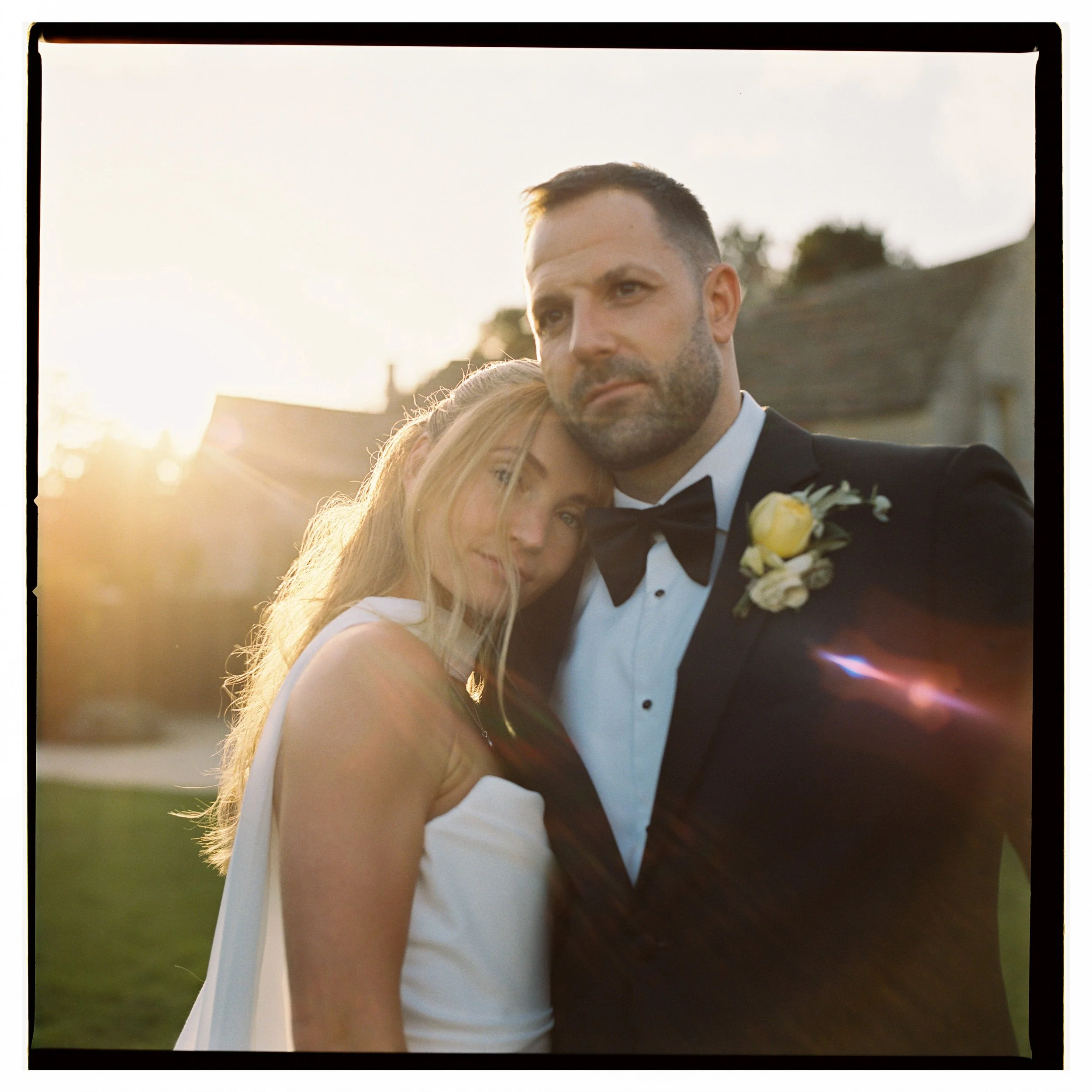 A bride and groom in wedding attire embracing outdoors at sunset, with the bride leaning her head on the groom's shoulder. The groom is wearing a black tuxedo with a boutonniere, and the bride is in a white wedding gown.