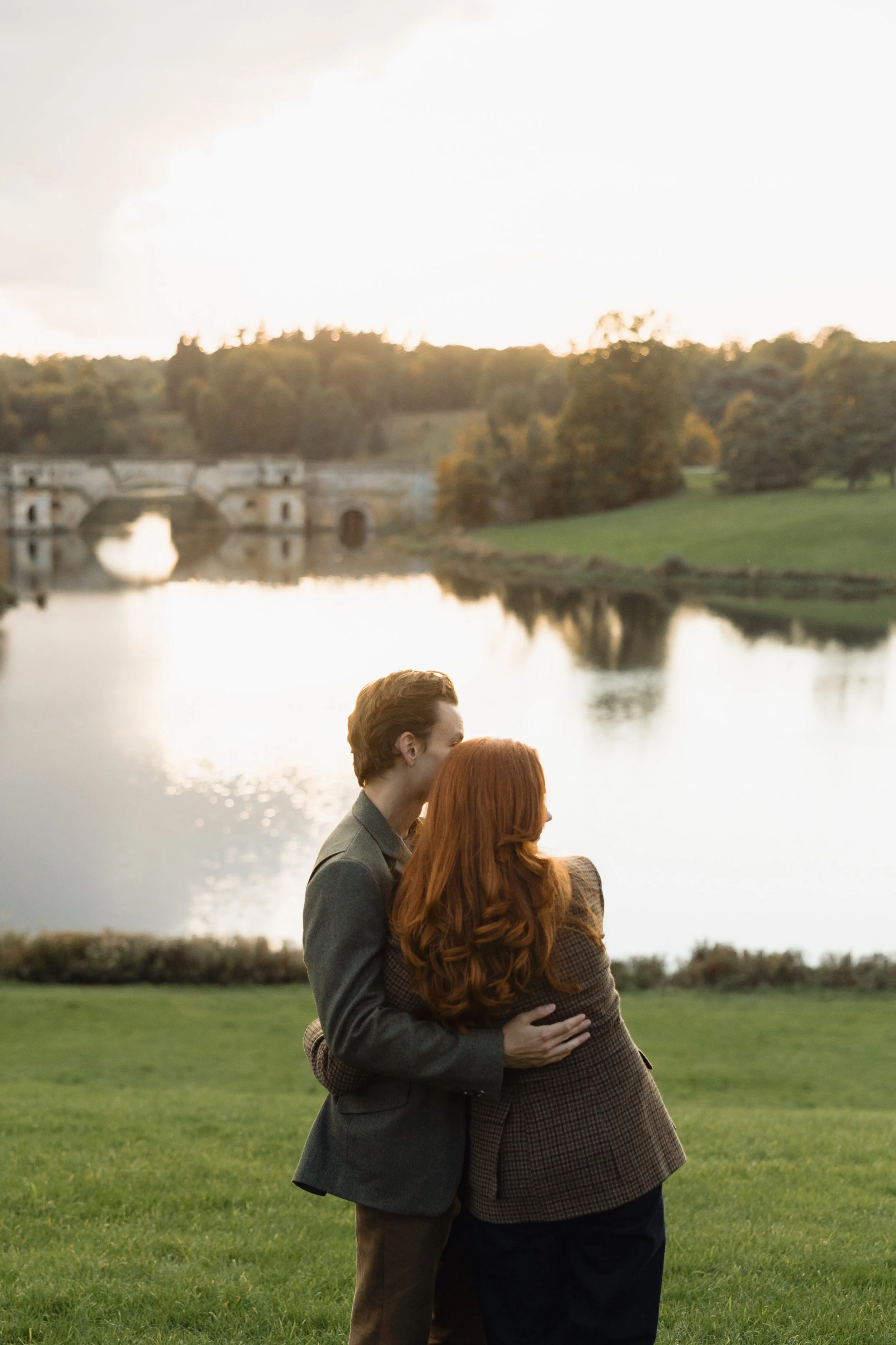 A couple with reddish hair embracing near a lake at sunset with trees and a stone bridge in the background.