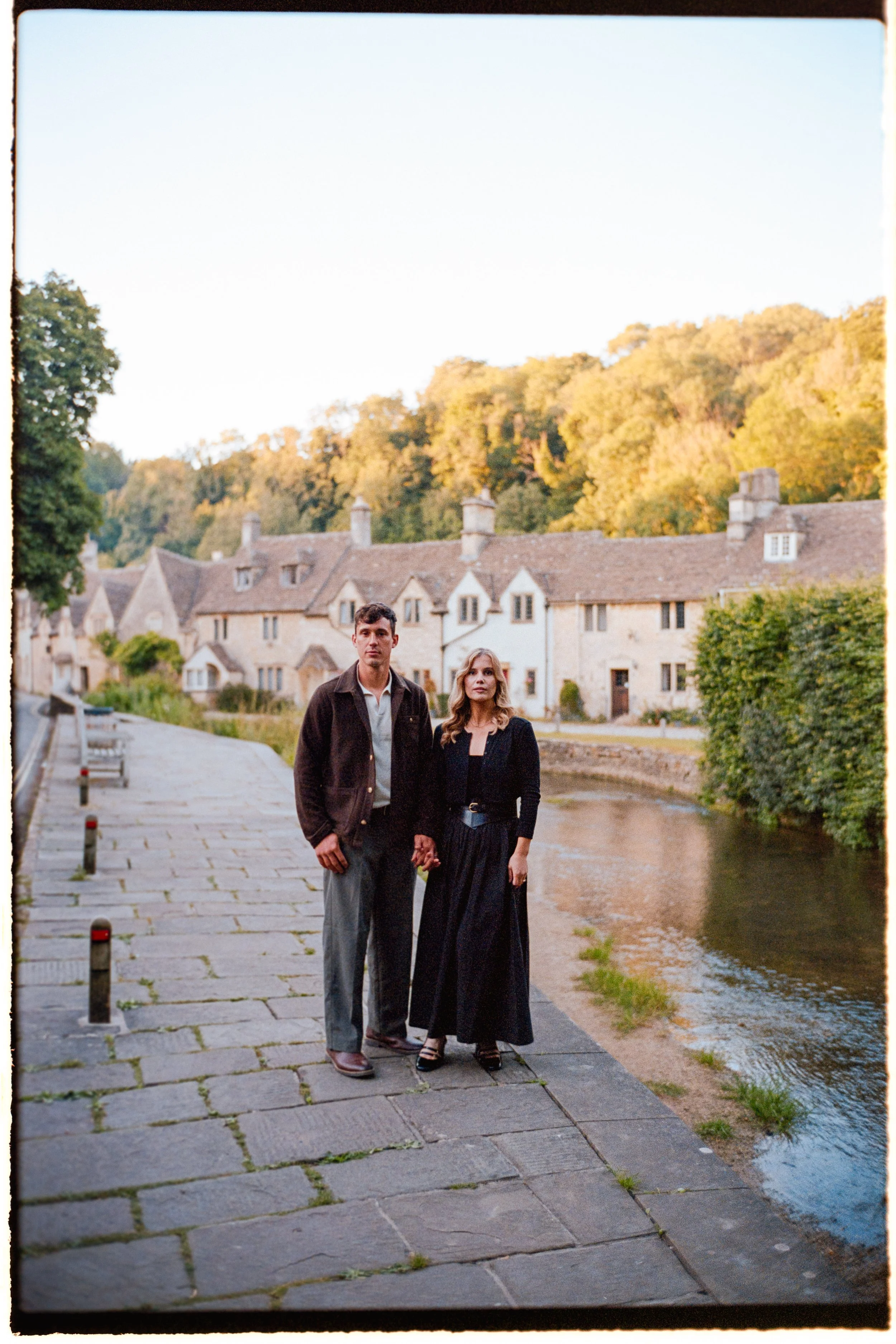A man and woman holding hands standing on a stone pathway beside a river, with an old village of stone houses and green trees in the background during sunset.
