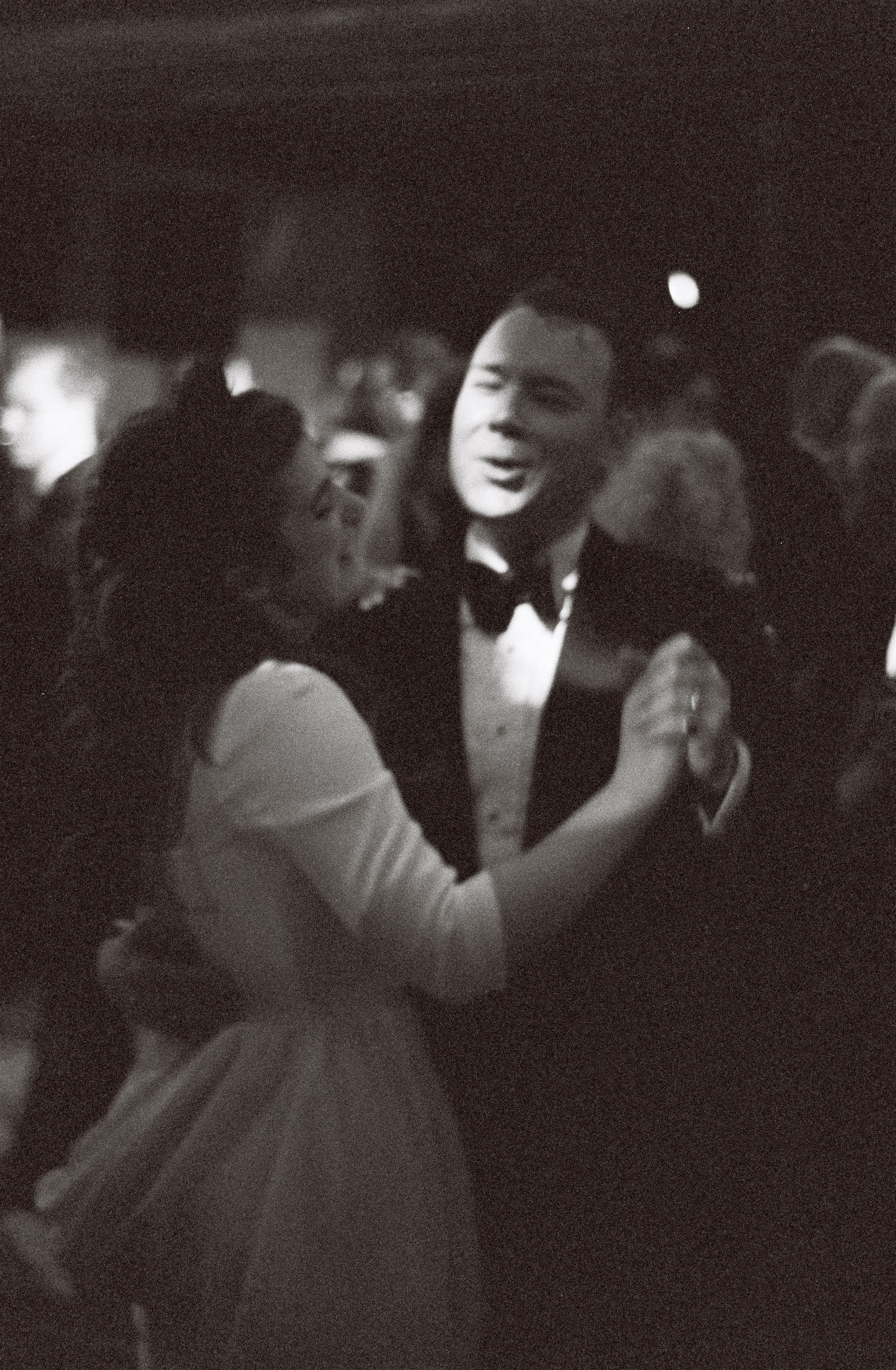 A black and white 35mm photo of a man and woman dancing closely at a formal event, both dressed in elegant attire, with other people dancing in the background.