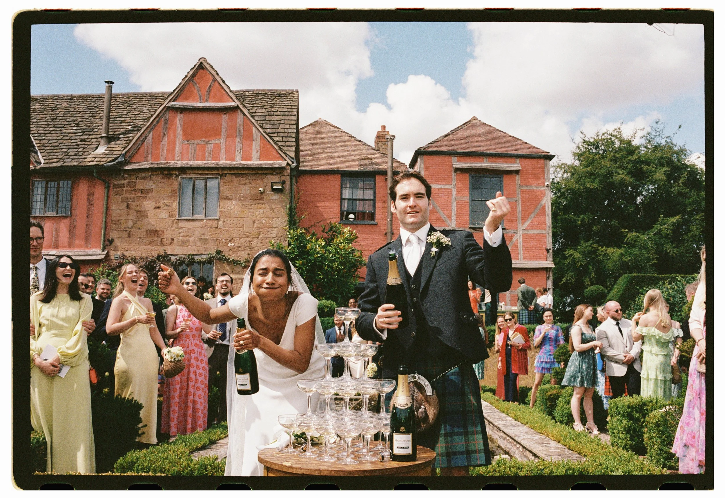 Wedding celebration outdoors with bride and groom raising champagne glasses, surrounded by guests, with a historic brick and stone building in the background.