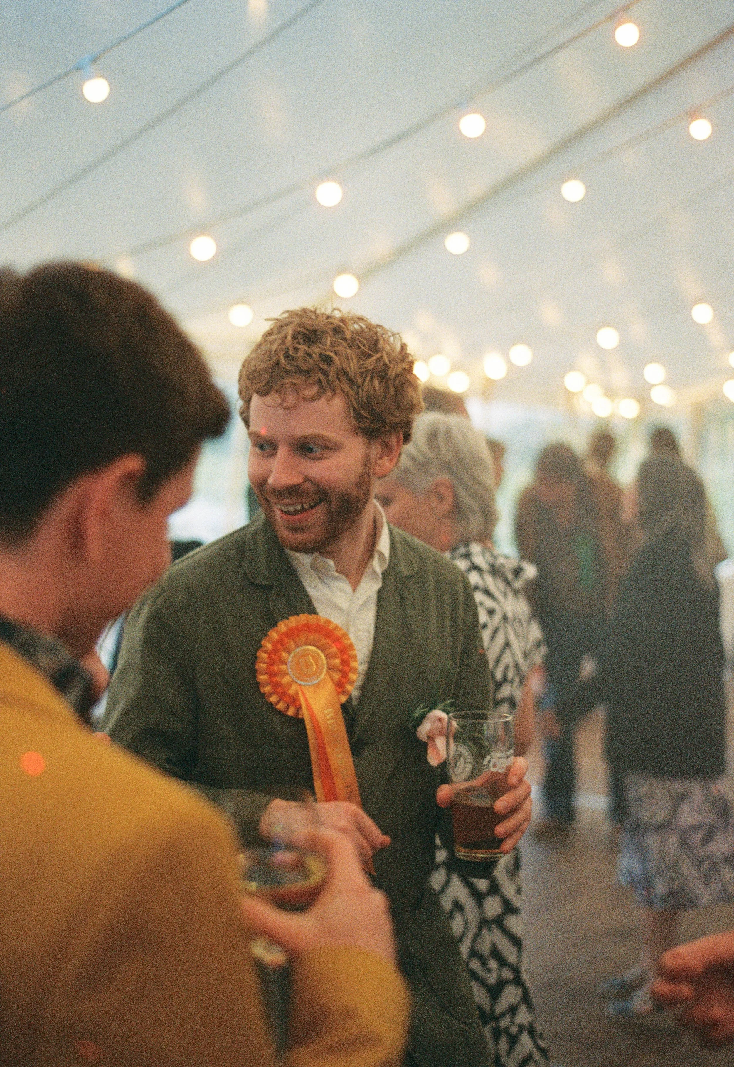 A man with curly hair and a beard, wearing a green jacket and a ribbon badge, holding a glass of beer, smiling and talking to a person in a yellow jacket at a lively indoor event with string lights.