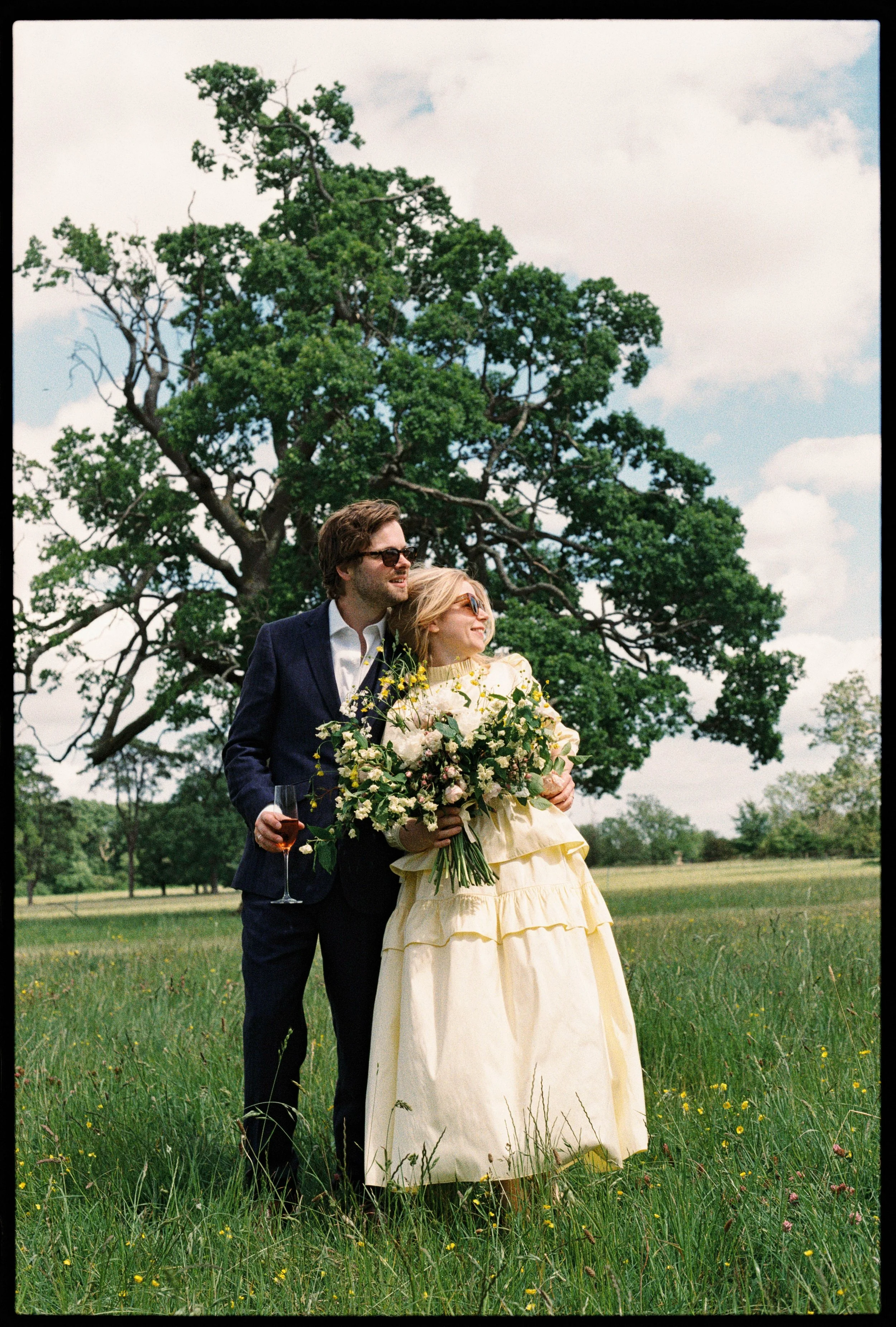 A couple standing outdoors on a grassy field with a large tree in the background. The woman is holding a bouquet of flowers and the man is holding a glass of wine. They are dressed in formal attire, with the woman in a cream-colored dress and the man